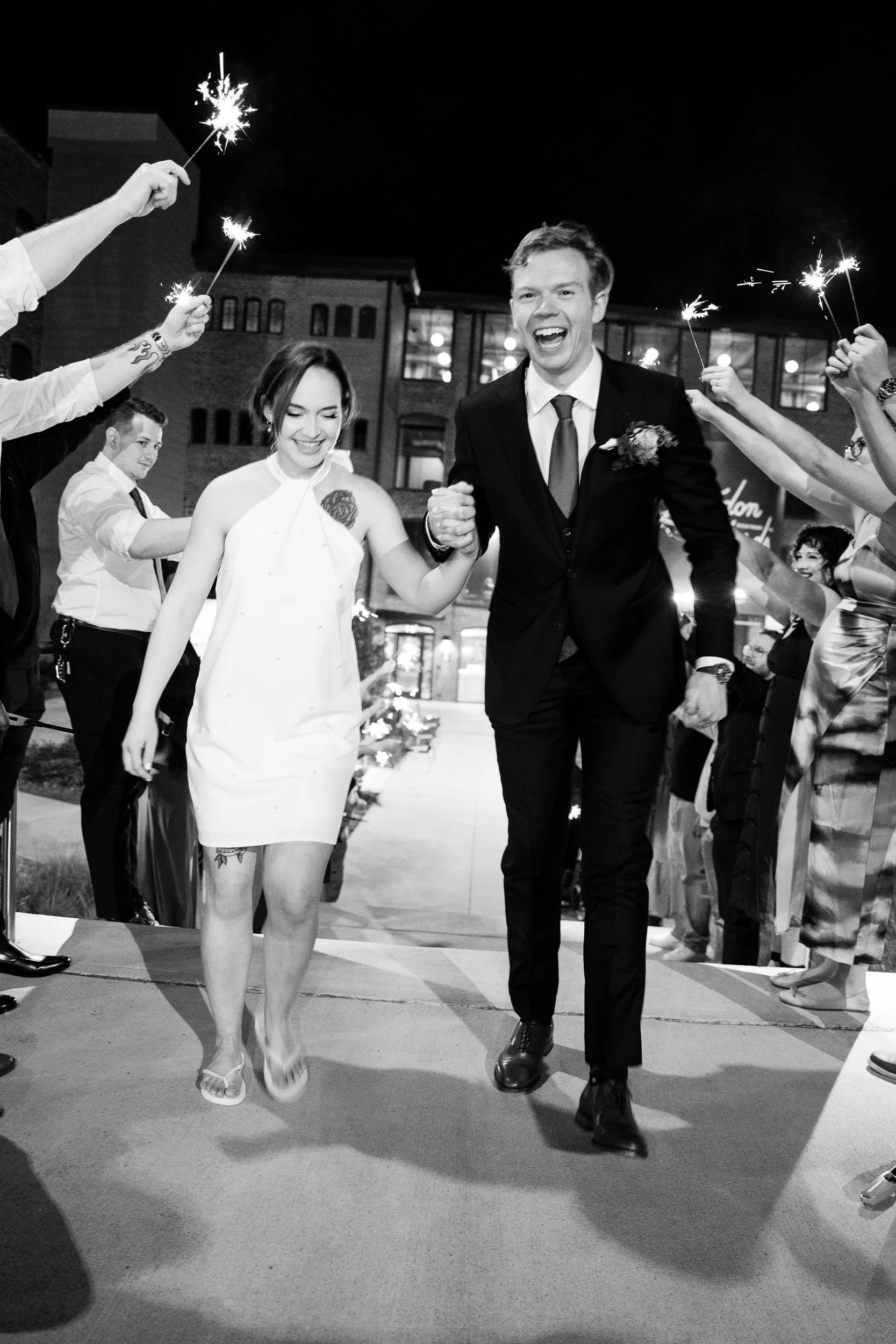 Black and white photo of a wedding couple walking through a sparklers parade at night, holding hands, smiling, with guests celebrating around them.