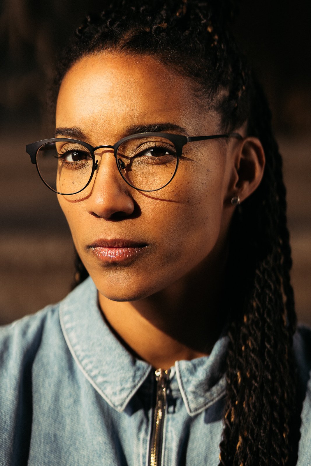 A close-up portrait of a woman with dark skin, wearing glasses and a denim jacket, with her hair styled in twists.
