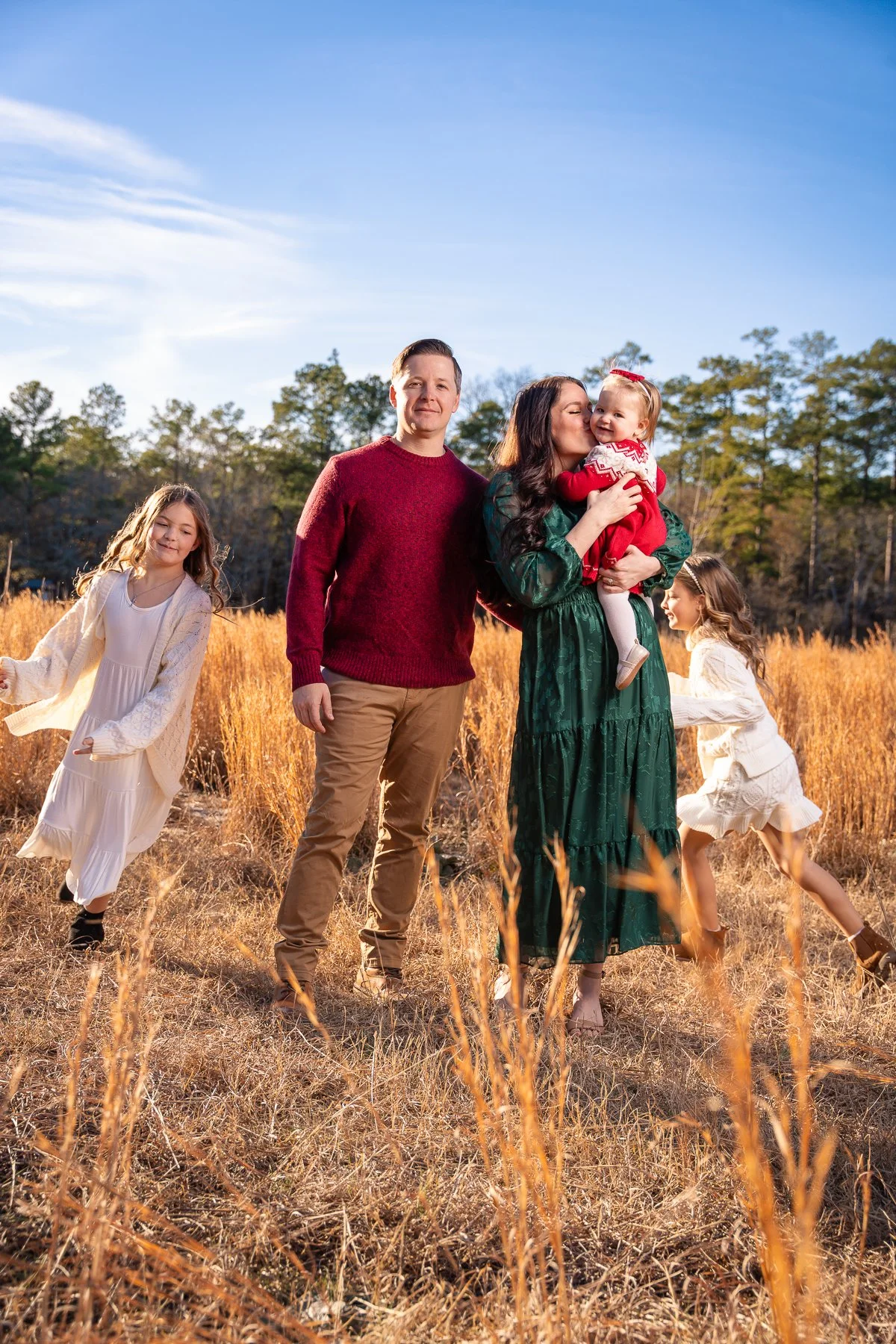 Family of five outdoors in a field during daytime, including a woman holding a young girl, a man standing beside her, and two girls playing by her sides.