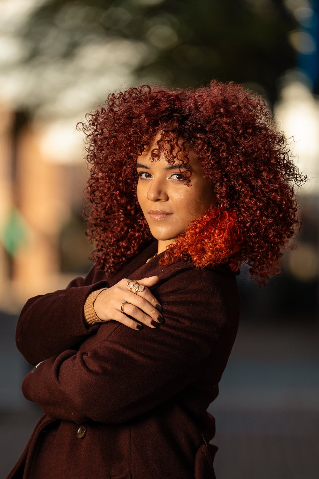 A woman with voluminous curly red hair looks at the camera, wearing a dark coat and rings, with her arms crossed.