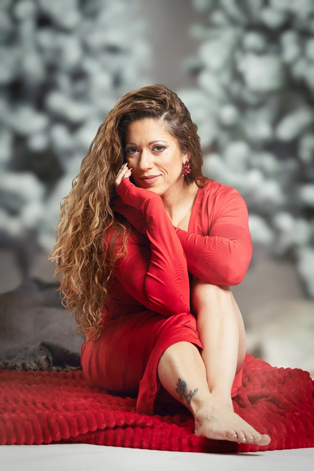 A woman in a red dress with curly hair sitting on a red blanket, posing with her chin resting on her hand, in front of a blurred background.