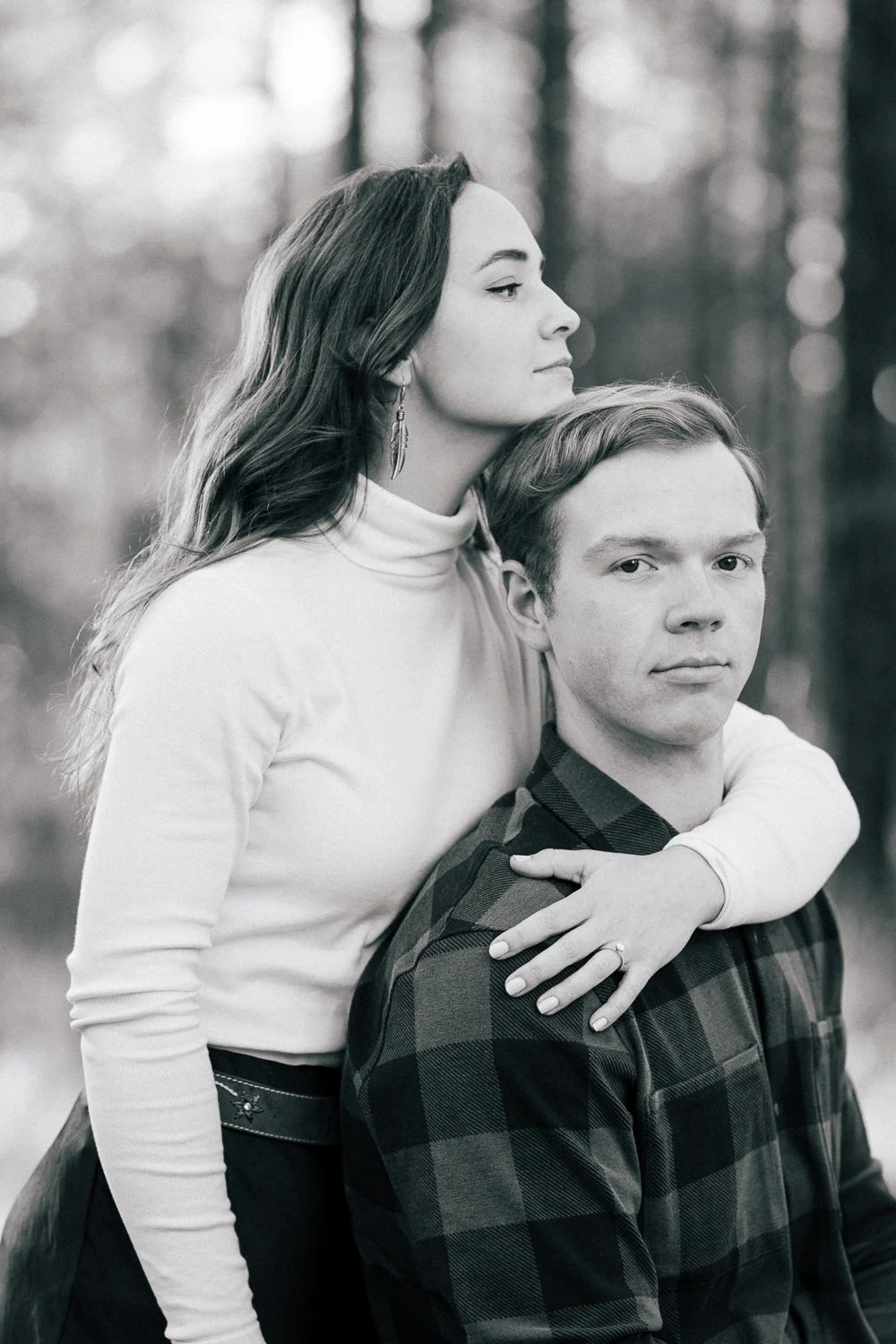 A black and white photo of a woman with long hair and a man. The woman is standing behind the man, resting her arms on his shoulders, and looking to the side. The man is sitting and has short hair, wearing a checkered shirt, looking directly at the c