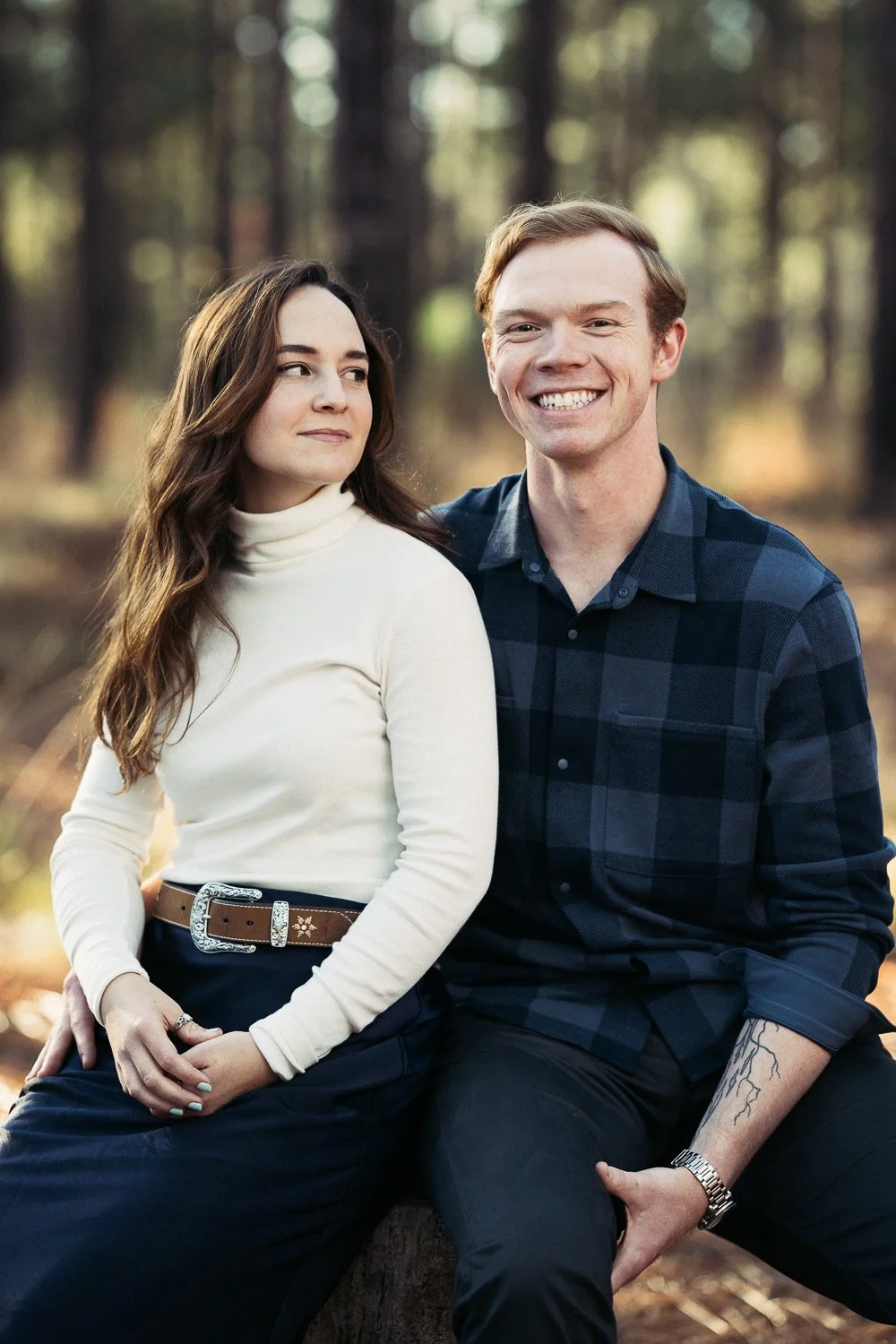 A woman and a man sitting outdoors in a forest, smiling and looking at each other.
