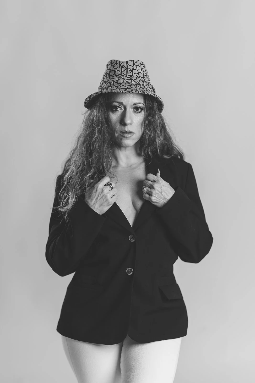 Black and white portrait of a woman with curly hair wearing a patterned bucket hat, a black blazer, and light-colored pants, with her hands adjusting her blazer.