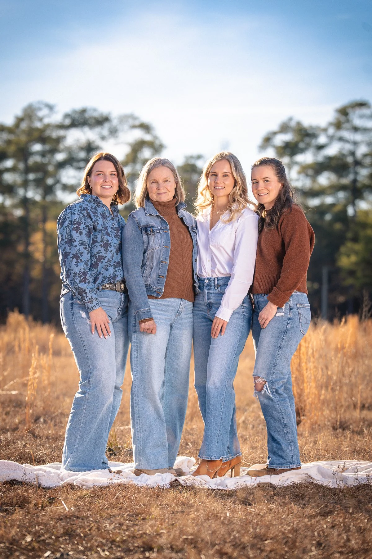 Four women standing outdoors in a grassy field during fall, smiling at the camera, dressed in casual denim and sweaters.