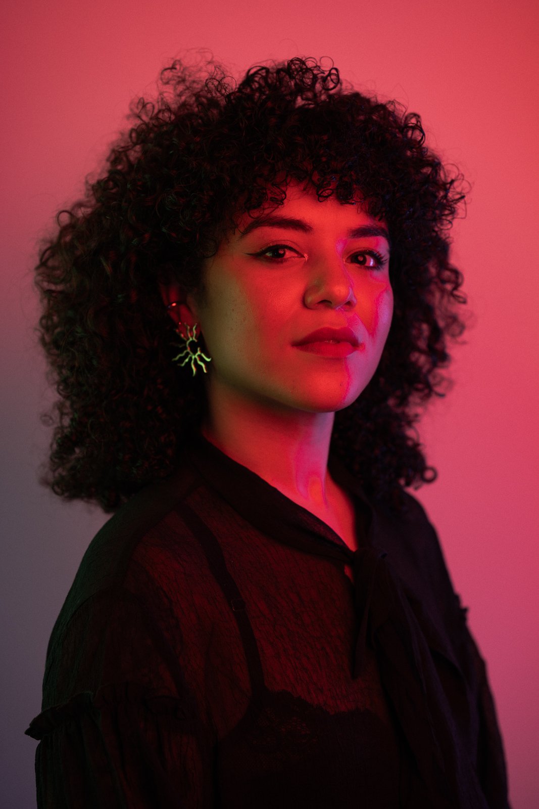 A woman with curly dark hair and light skin poses against a pink and purple gradient background, wearing earrings shaped like branches and a black sheer top.