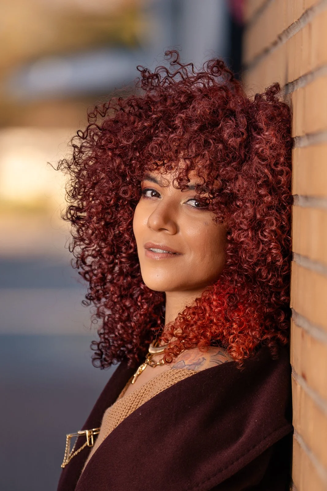 A woman with curly red hair and light skin leaning against a brick wall outdoors during golden hour.