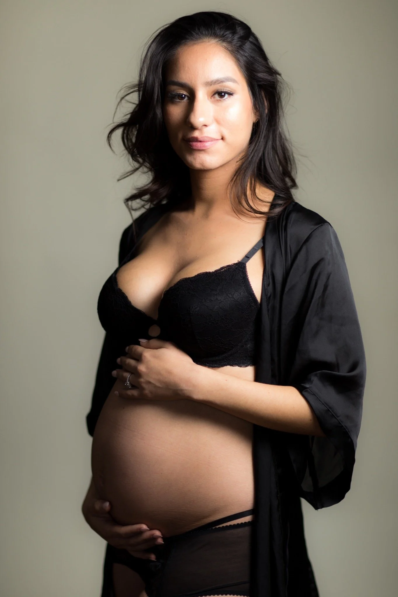 Portrait of a pregnant woman with black hair in black lingerie and a satin robe, standing against a plain background.