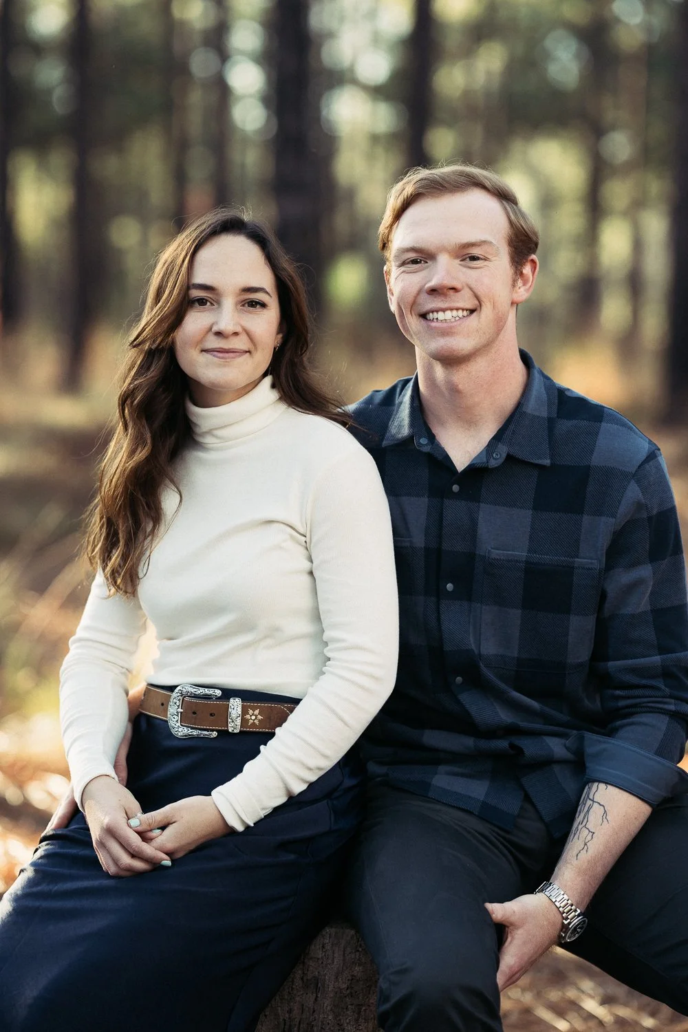 A young woman with long brown hair and a young man with short blonde hair sitting outdoors in a forest, smiling at the camera.