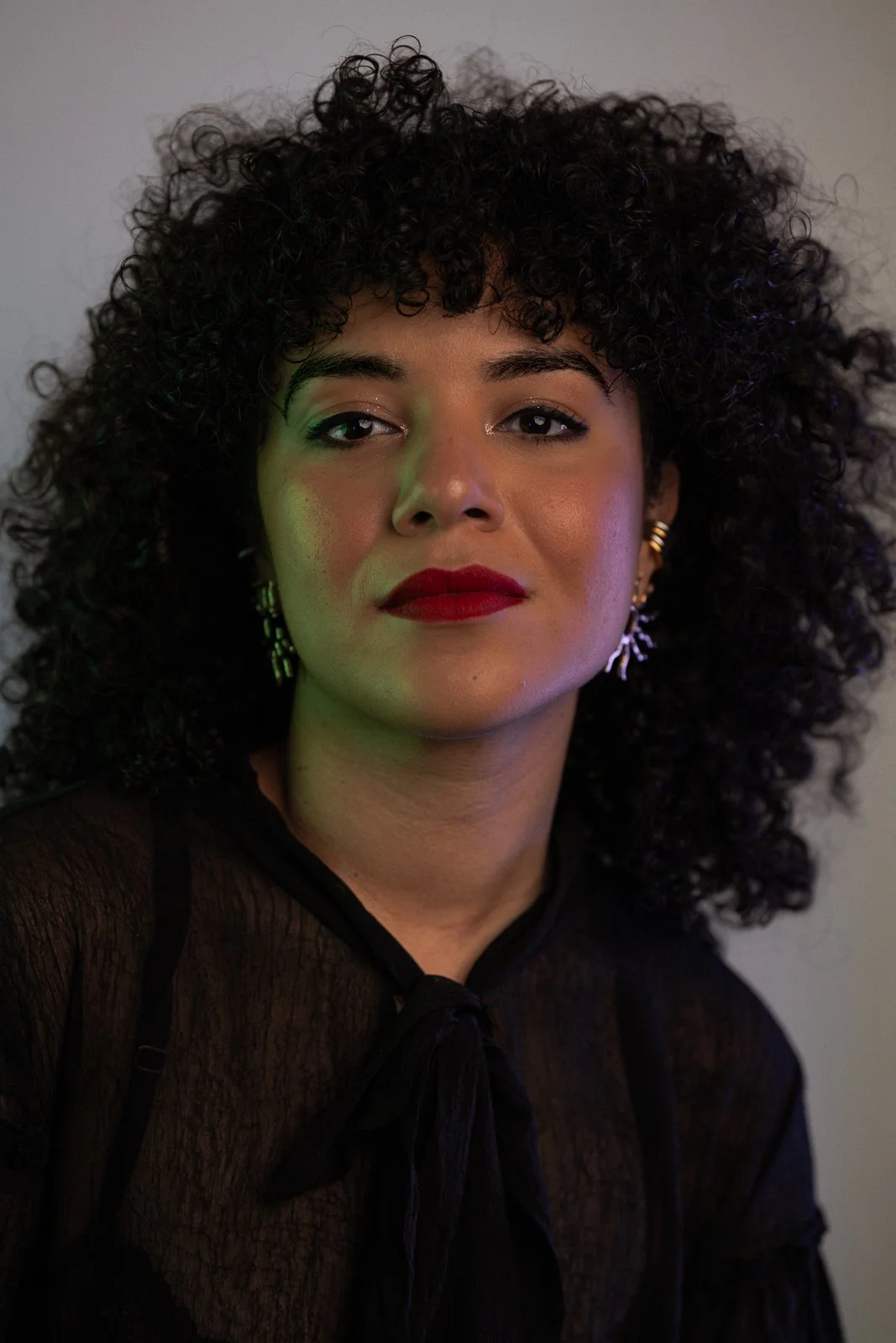 A woman with curly black hair wearing red lipstick and earrings, dressed in a black sheer top with a bow at the neckline, posing against a plain wall.