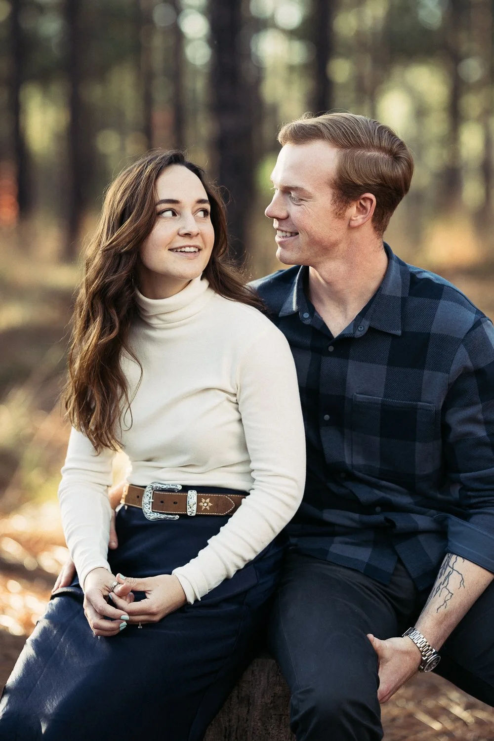 A young woman and a young man sitting on a log in a forest, looking at each other and smiling. The woman has long brown hair and is wearing a cream turtleneck and dark pants. The man has light brown hair, is wearing a blue checkered shirt, and has a 