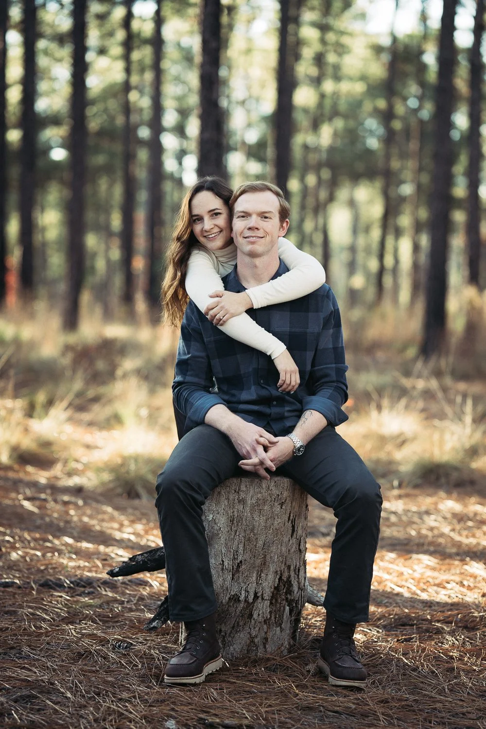 A young couple sitting on a tree stump in a pine forest, with the woman embracing the man from behind and both smiling.