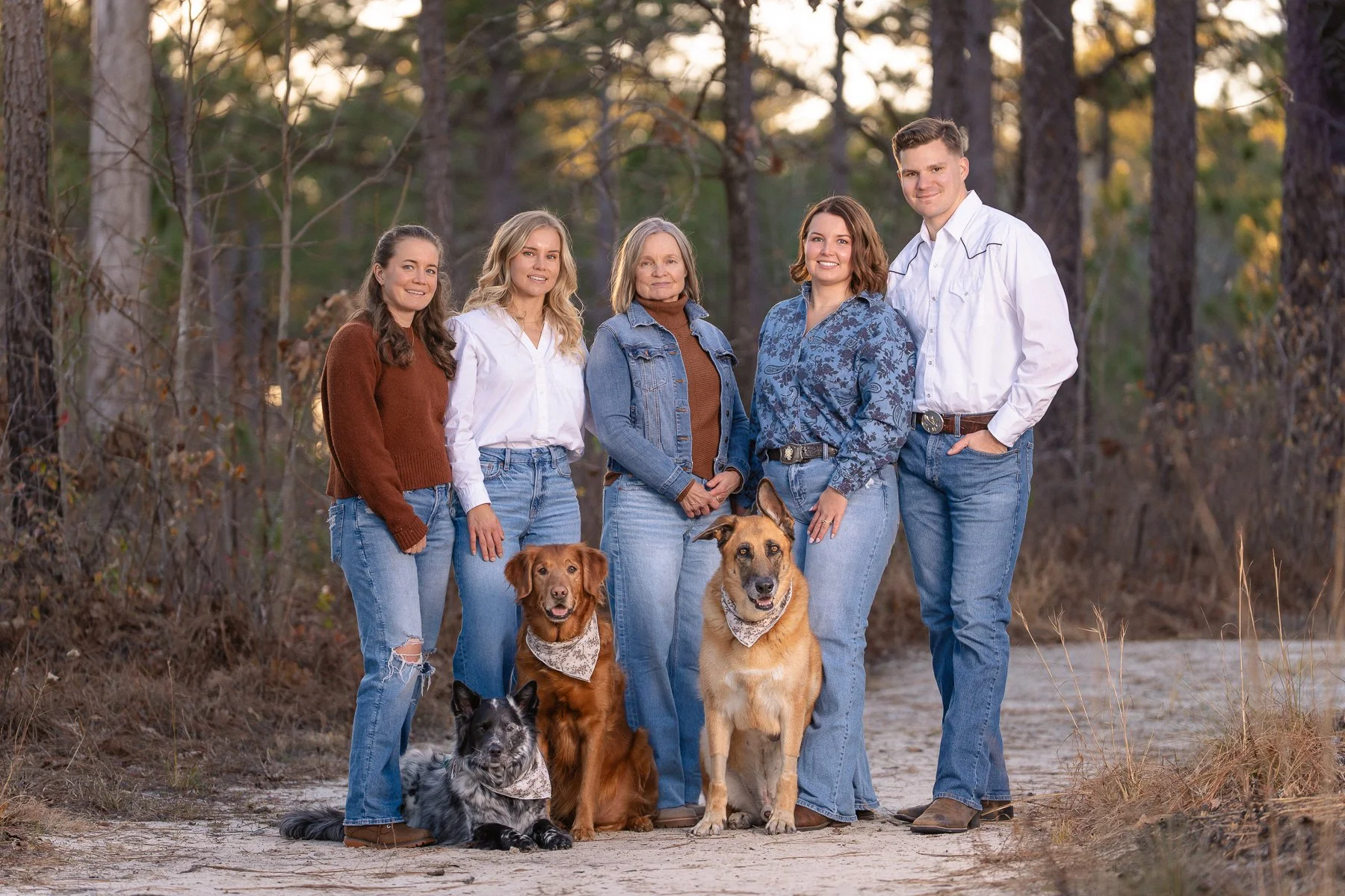 A group of five women and one man standing outdoors in a wooded area with three dogs sitting in front of them. The women are dressed casually, with some wearing jeans and long-sleeve shirts or sweaters, and the man is wearing a white shirt and jeans.