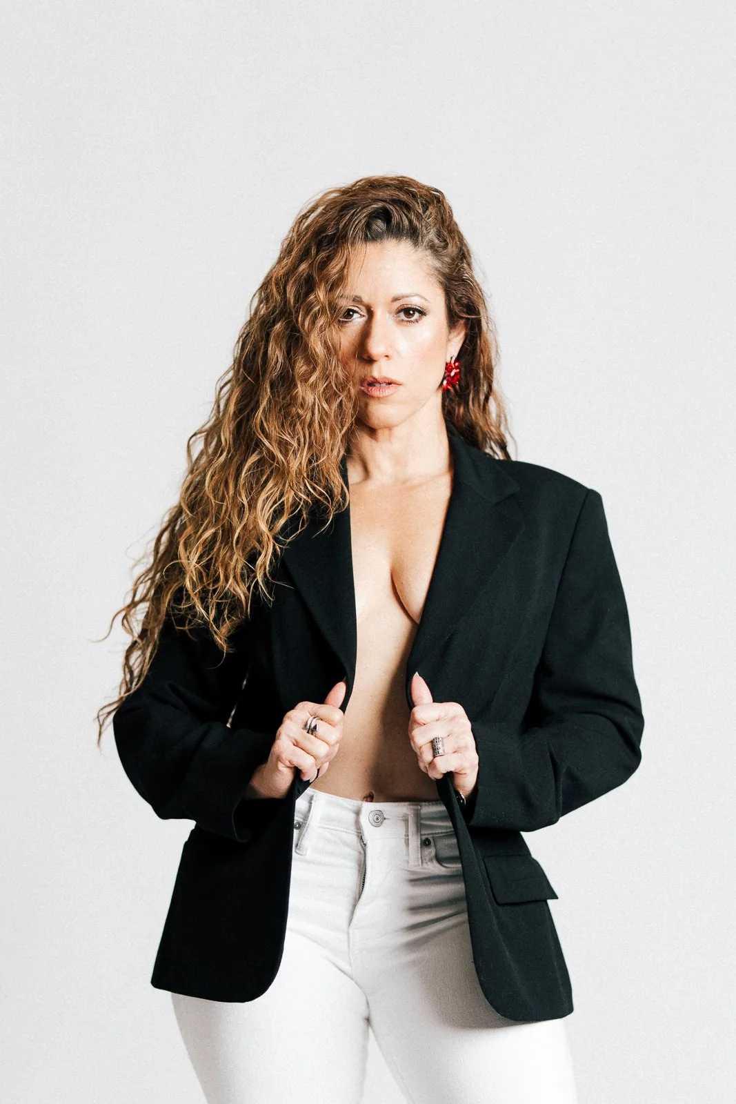 Woman with long curly hair wearing a black blazer and white pants, holding the blazer open with both hands, against a plain white background.