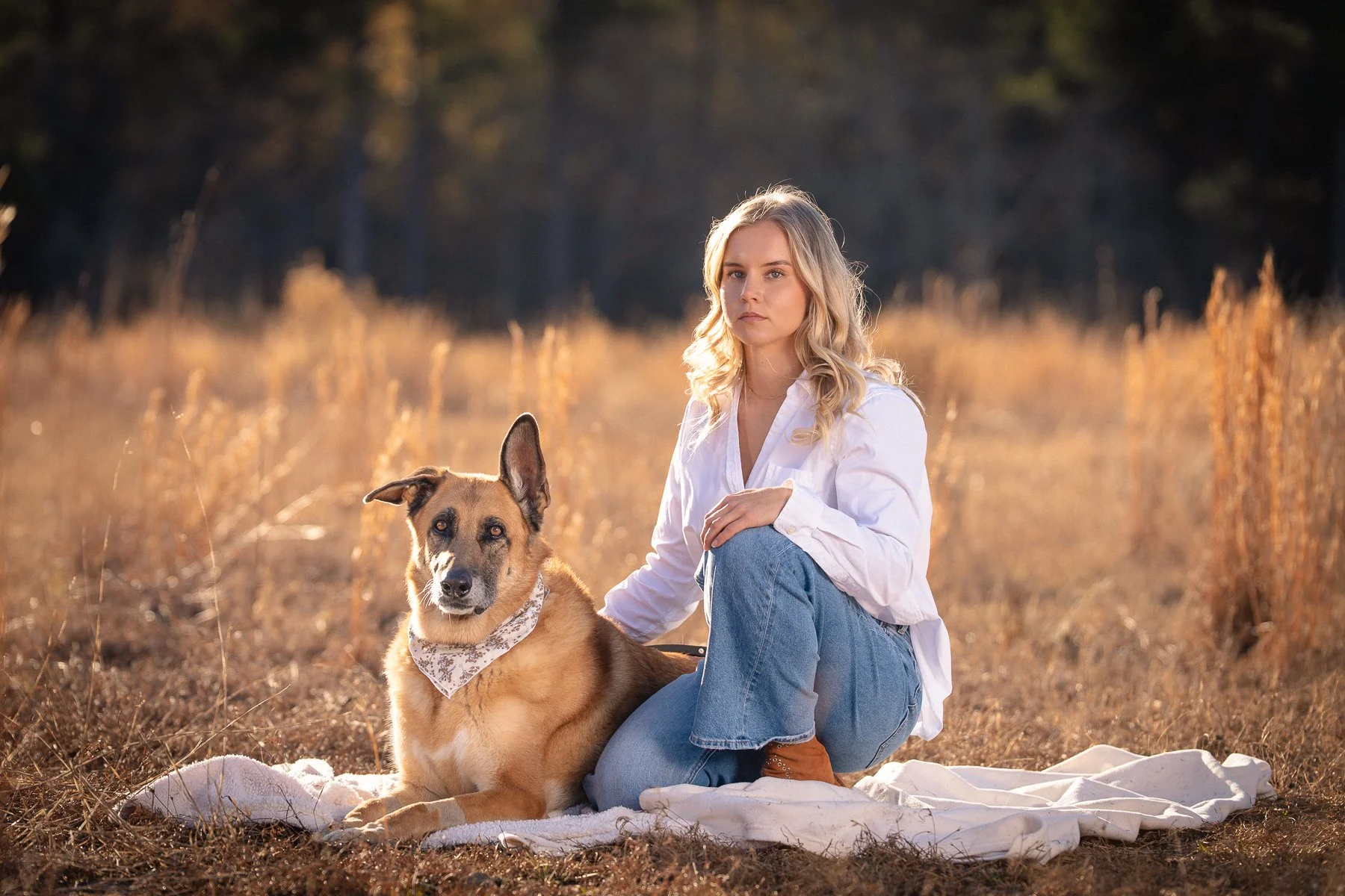 A young woman sitting on a blanket in a field with a dog, both looking at the camera. The woman has blonde hair and is wearing a white shirt and blue jeans. The dog is a mixed breed with a bandana around its neck.