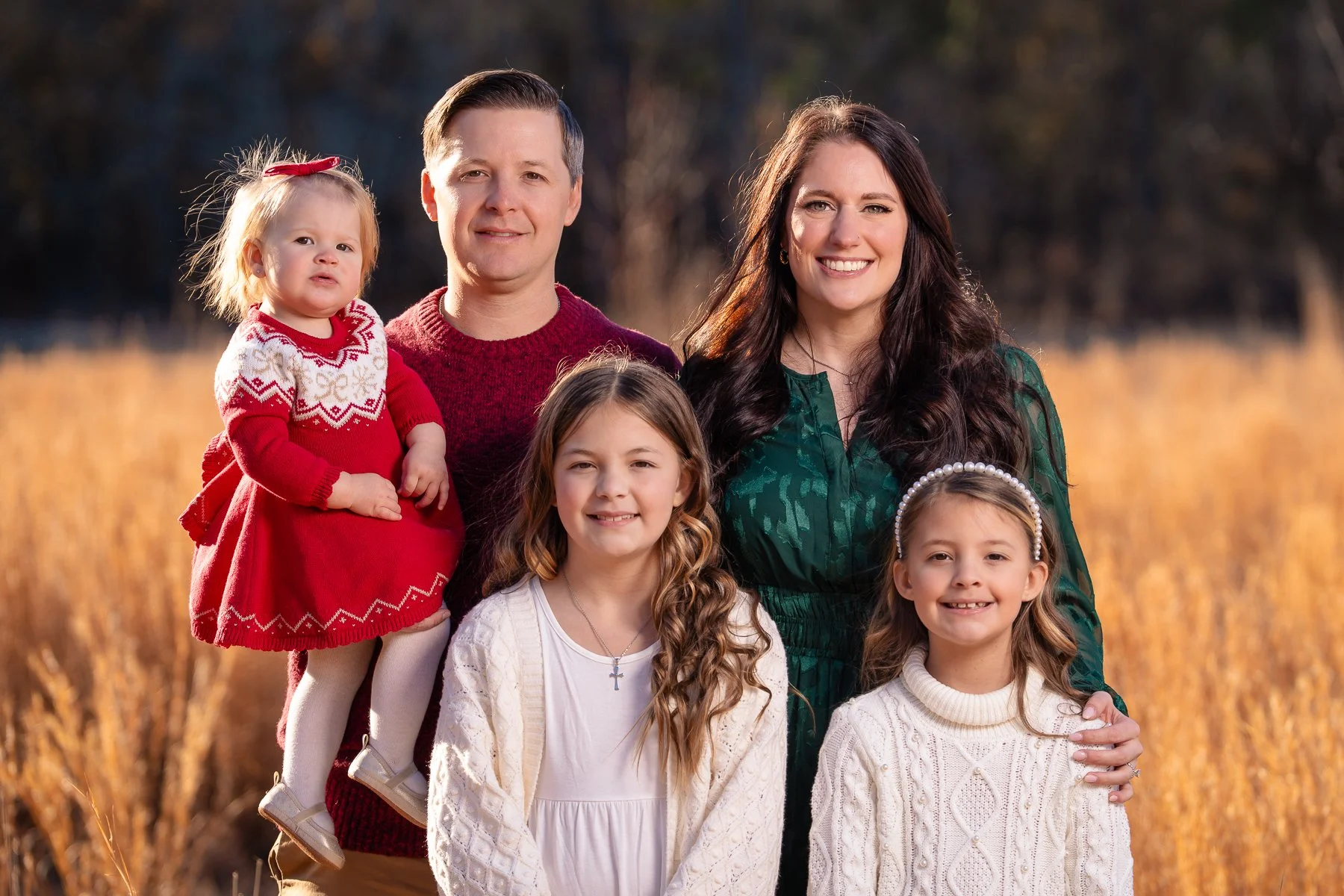 Family of five posing outdoors in fall, including father, mother, and three daughters, with golden field and trees in background.