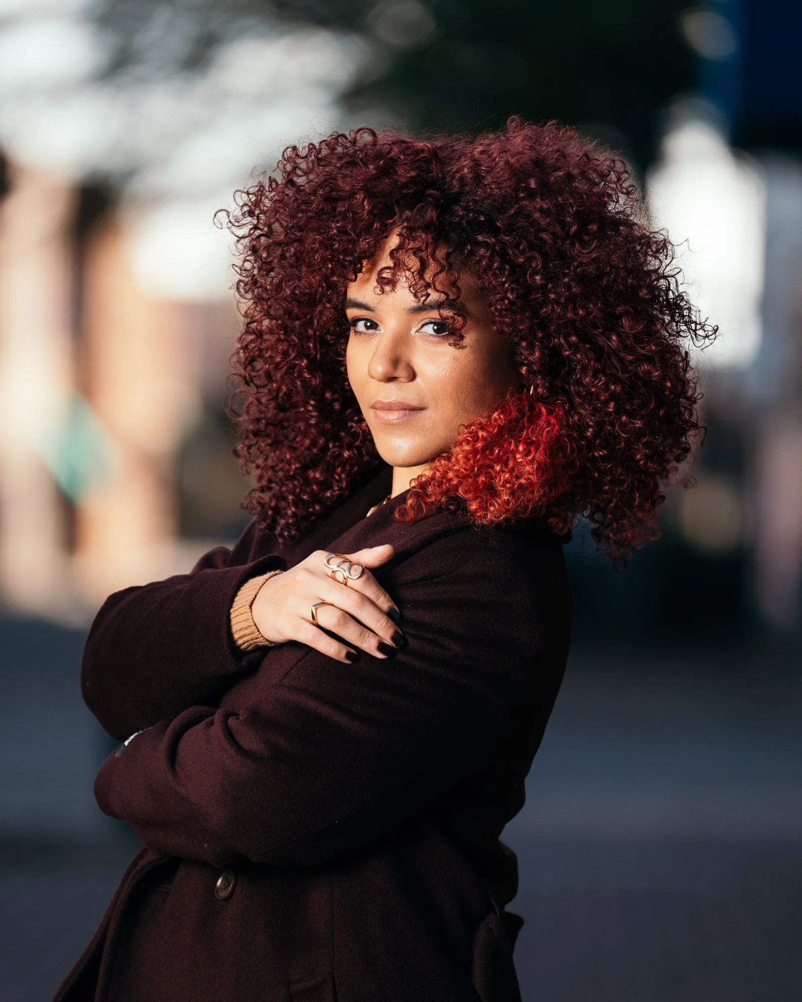 A woman with curly red hair, wearing a dark coat and rings, standing outdoors in daylight with blurred background.