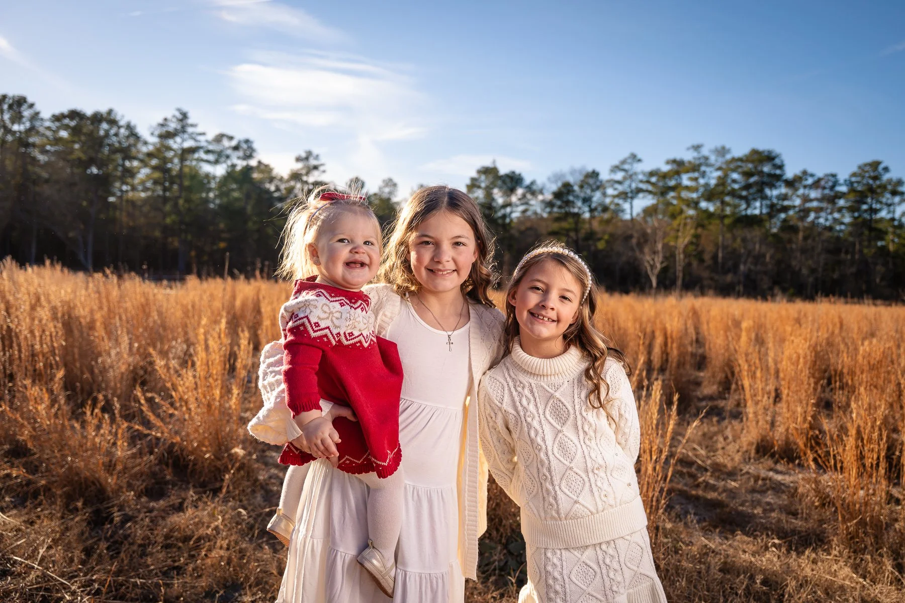 Three young girls smiling outdoors in a field of tall, golden grasses with trees and a blue sky in the background.