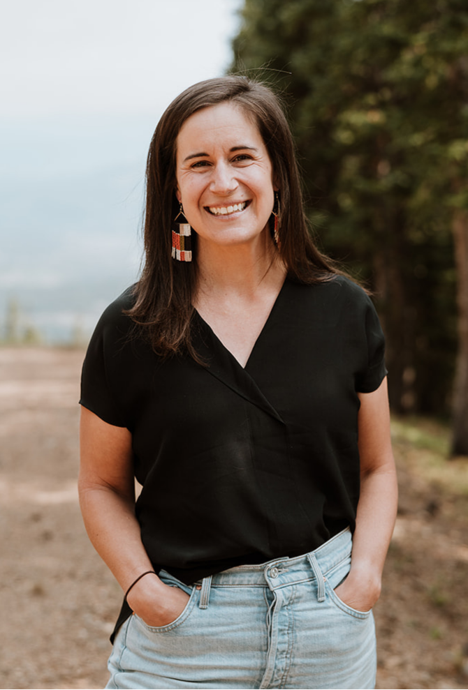 Carrie Andrews smiling outdoors in front of trees and a cloudy sky, wearing a black top, faded jeans, and colorful earrings.