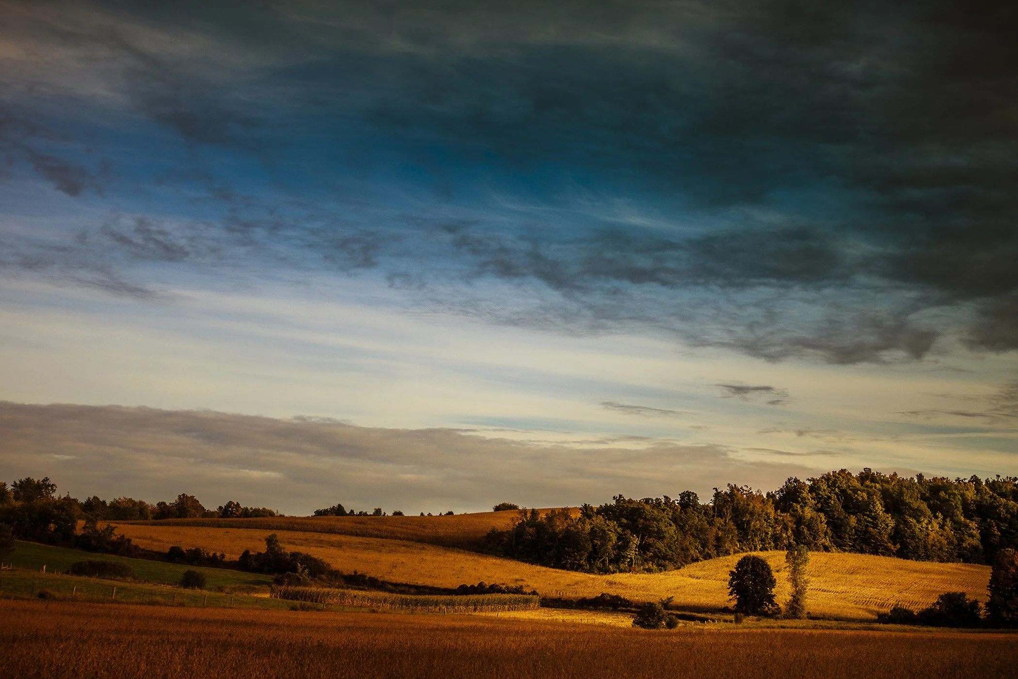 Farmland with rolling hills, trees, and a colorful sky with clouds in early evening light.