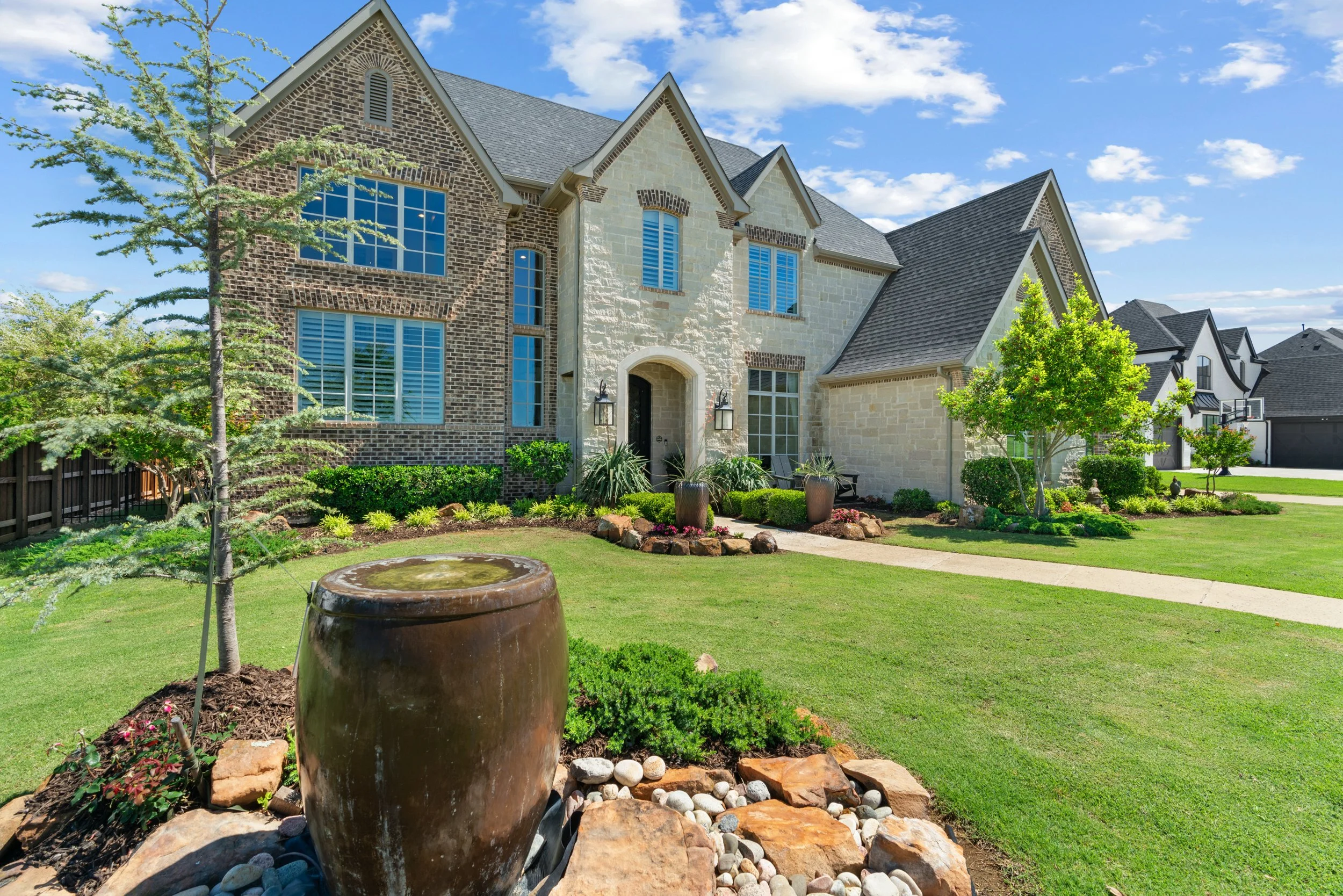 Front yard of a large, modern house with a well-maintained lawn, landscaping, and a water fountain feature in the foreground, under a partly cloudy sky.