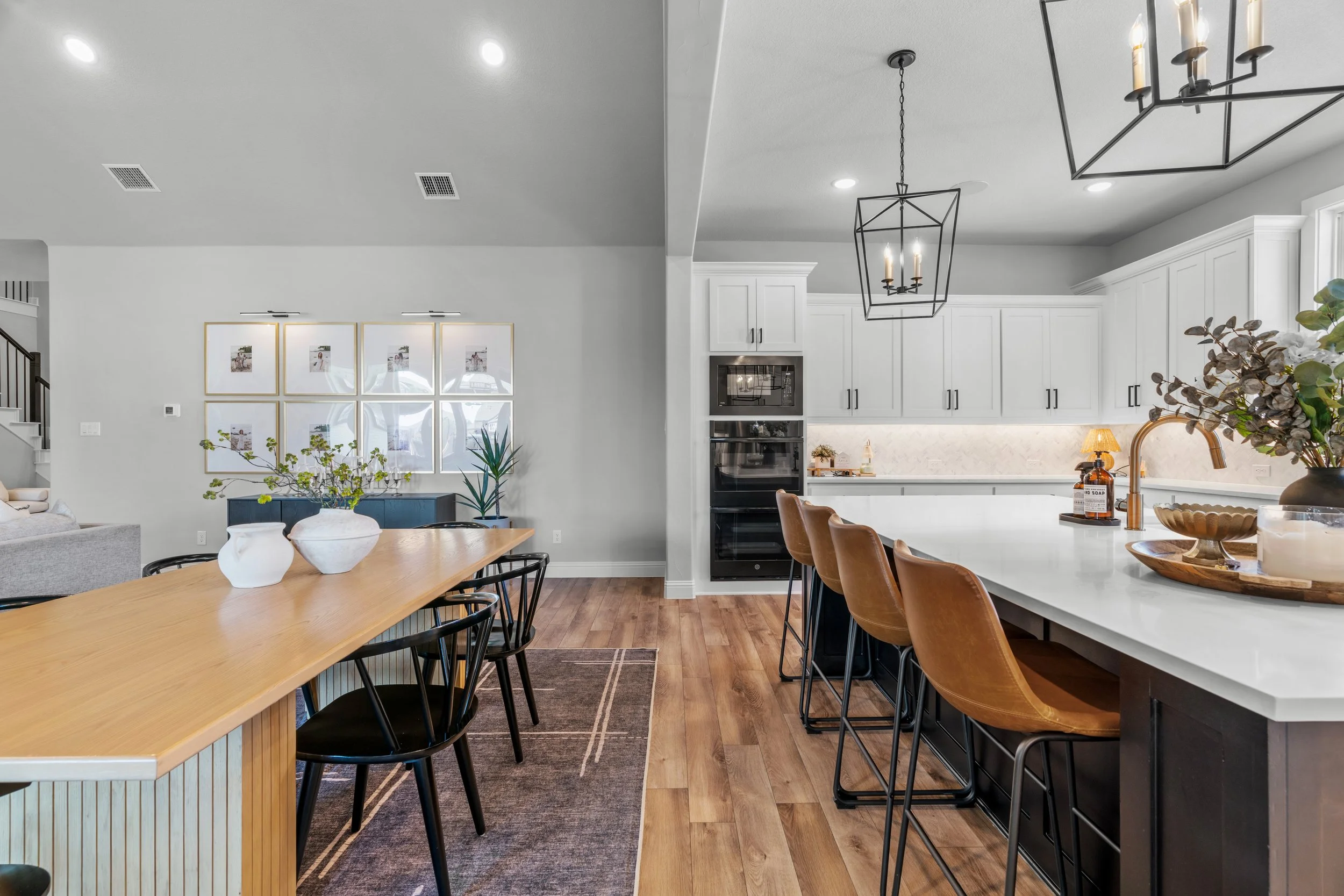Modern open-concept kitchen and dining area with wood flooring, white cabinets, a large white island with brown leather bar stools, black pendant lights, and a dining table with black chairs and white vases with green foliage.