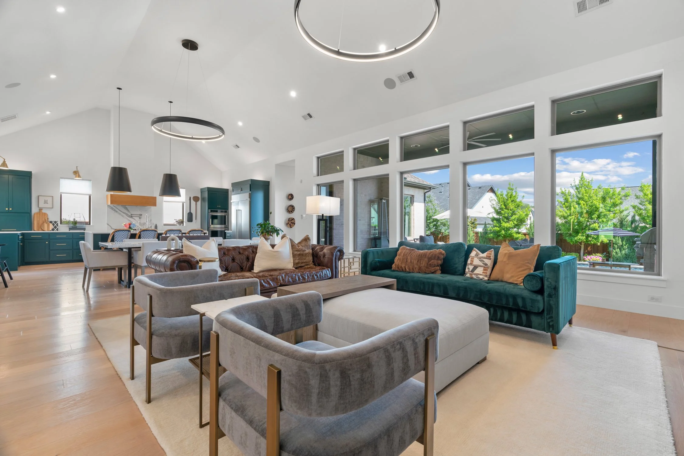 Spacious, modern open-plan living room with large windows offering a garden view, featuring various colorful sofas, a wooden dining table, teal kitchen cabinets, pendant lighting, and a light wood floor.