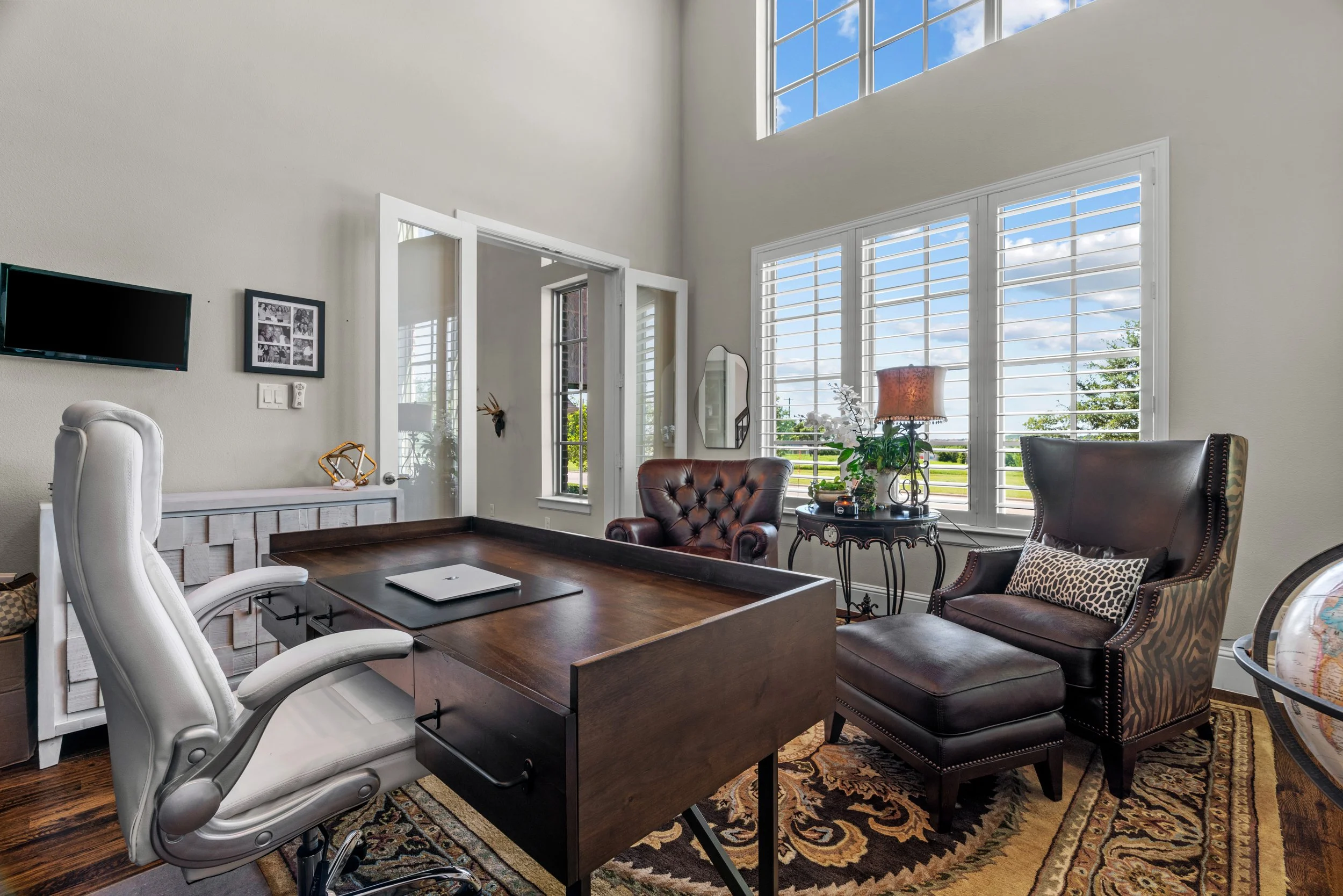 Home office with a large wooden desk, white leather chair, black leather armchair with ottoman, large windows with white shutters, potted plants, and decorative rug.