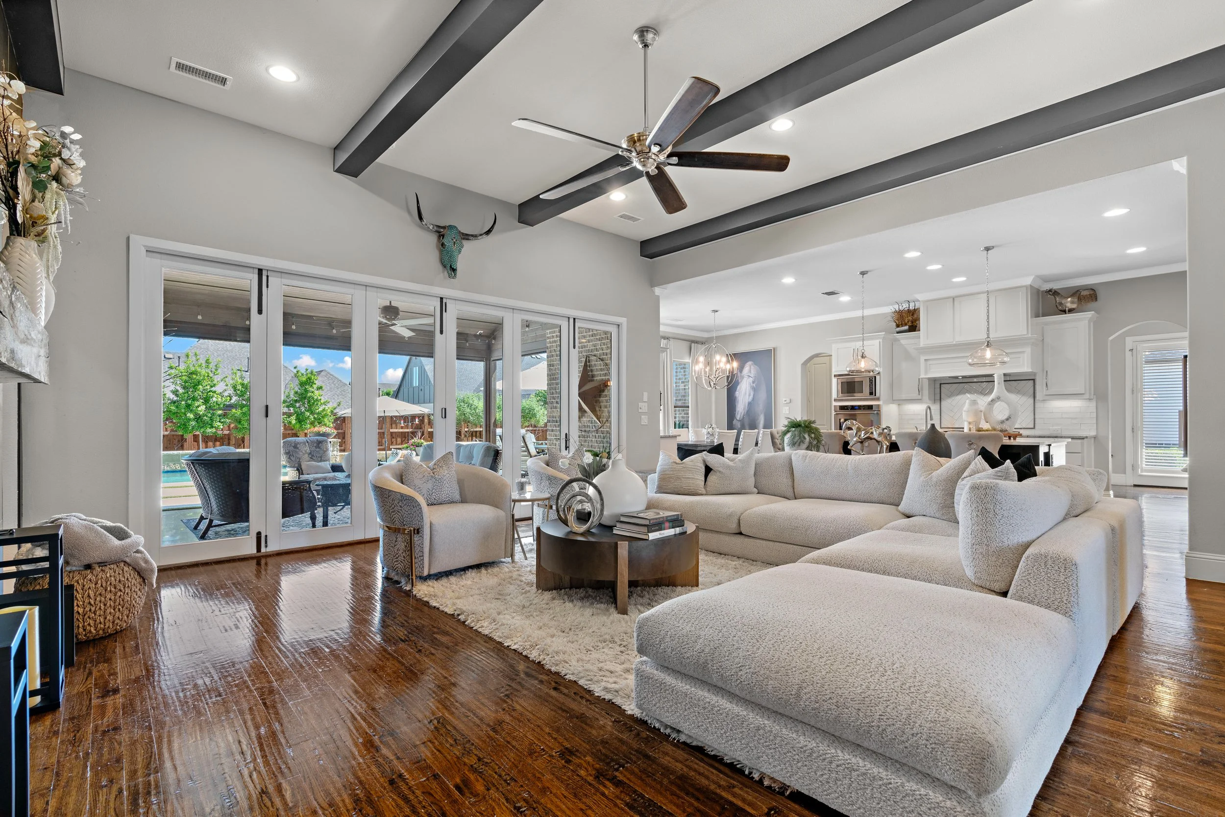 Bright living room with white sectional sofa, beige armchair, and a round coffee table on a plush rug, with sliding glass doors leading to an outdoor patio and pool area.