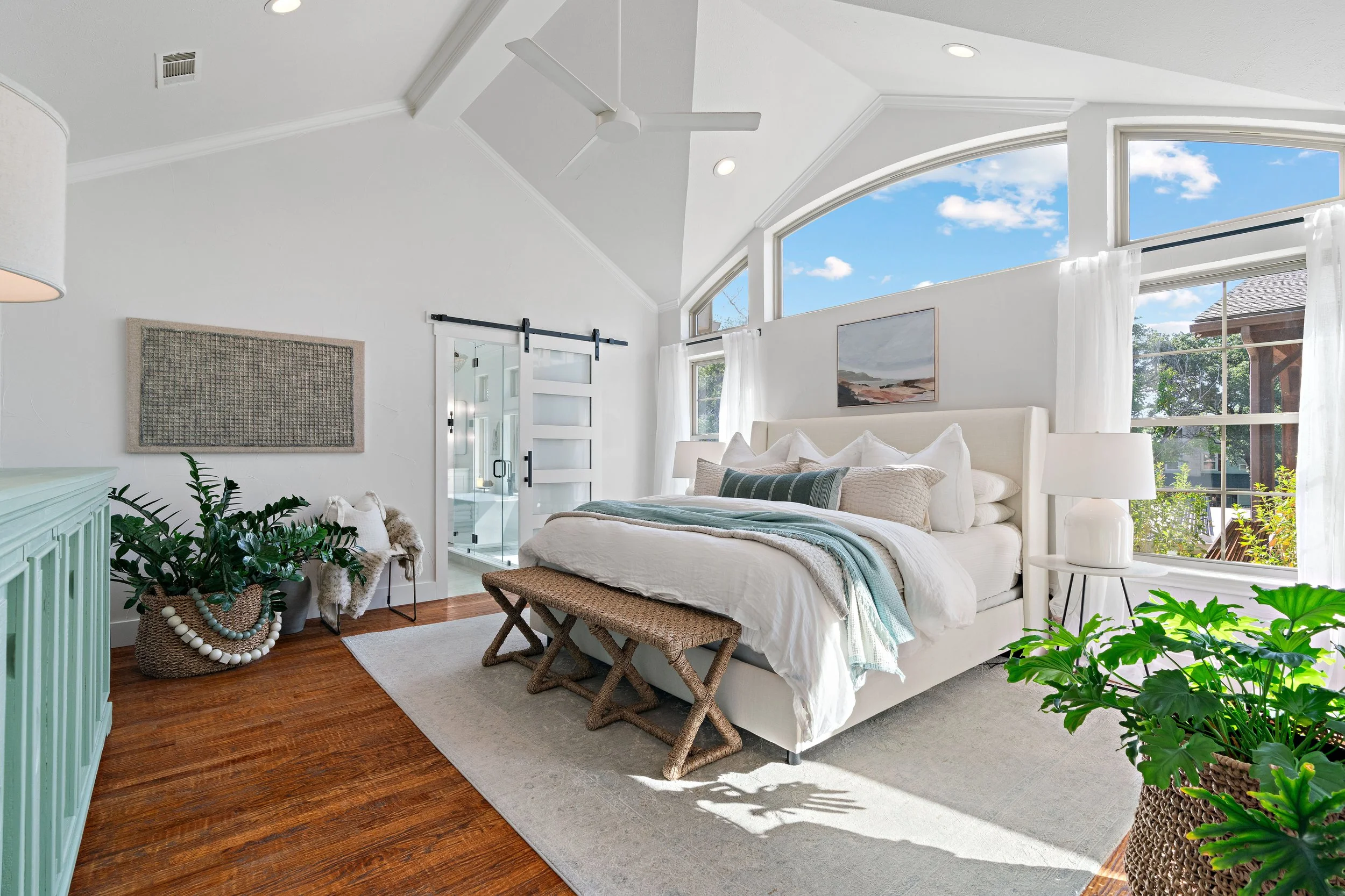 Bright bedroom with white walls, large windows showing a blue sky, a white bed with multiple decorative pillows, a small side table with a lamp, potted plants, and a sliding glass door leading to a bathroom.