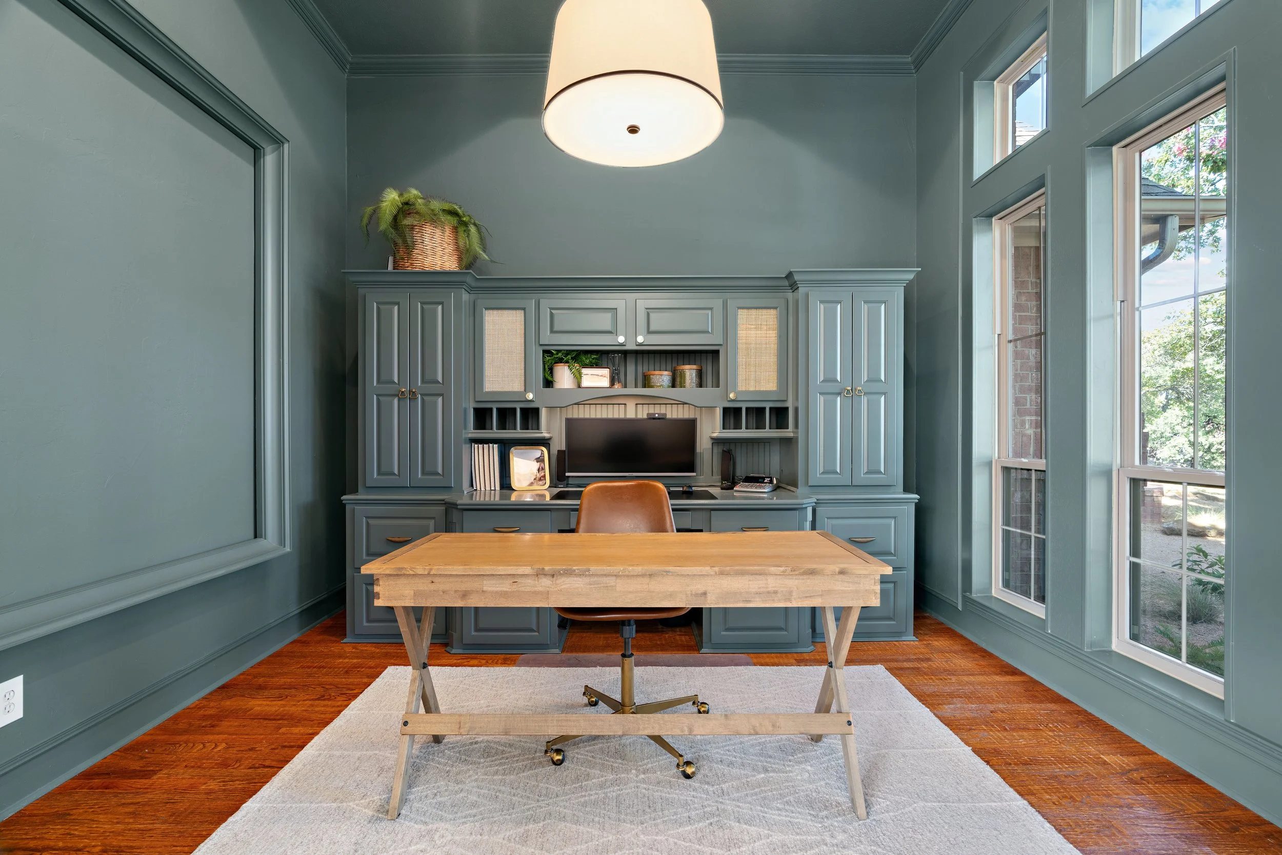 Home office with blue built-in cabinetry, wooden desk, brown office chair, and large windows showing outdoor greenery, illuminated by a ceiling light.