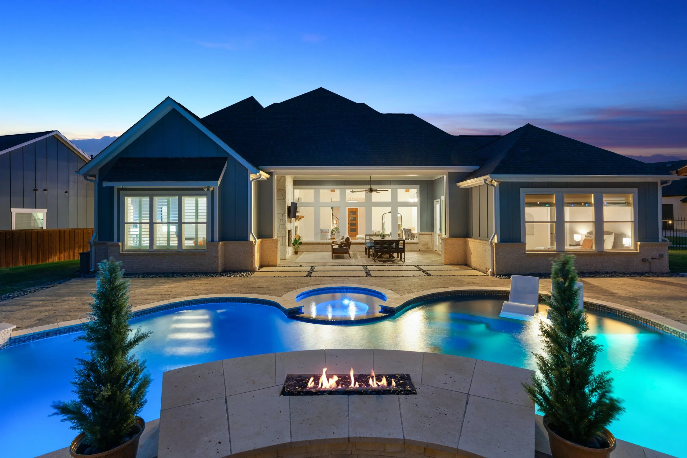 A modern house with a backyard pool at dusk, featuring a built-in fire pit and outdoor furniture in the patio area.