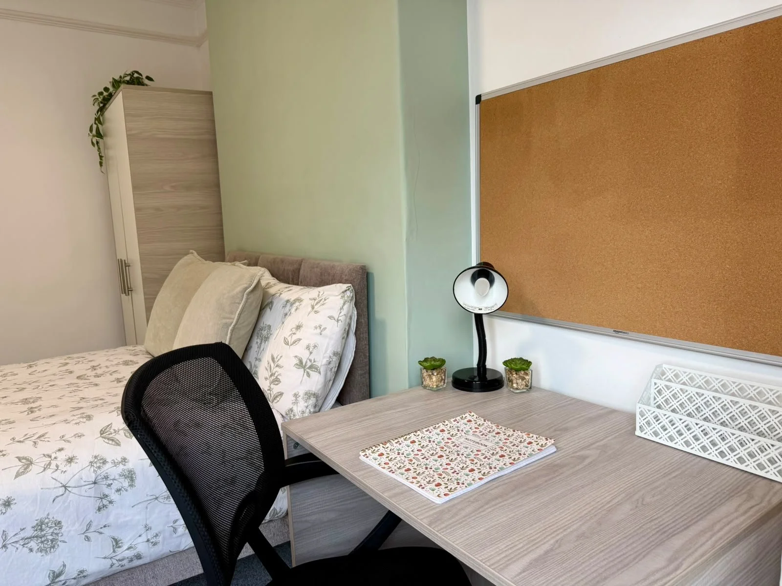 A bedroom and workspace setup with a bed, pillows, a desk, a black mesh chair, a lamp, two small plants, a corkboard, and a notebook with a floral cover.