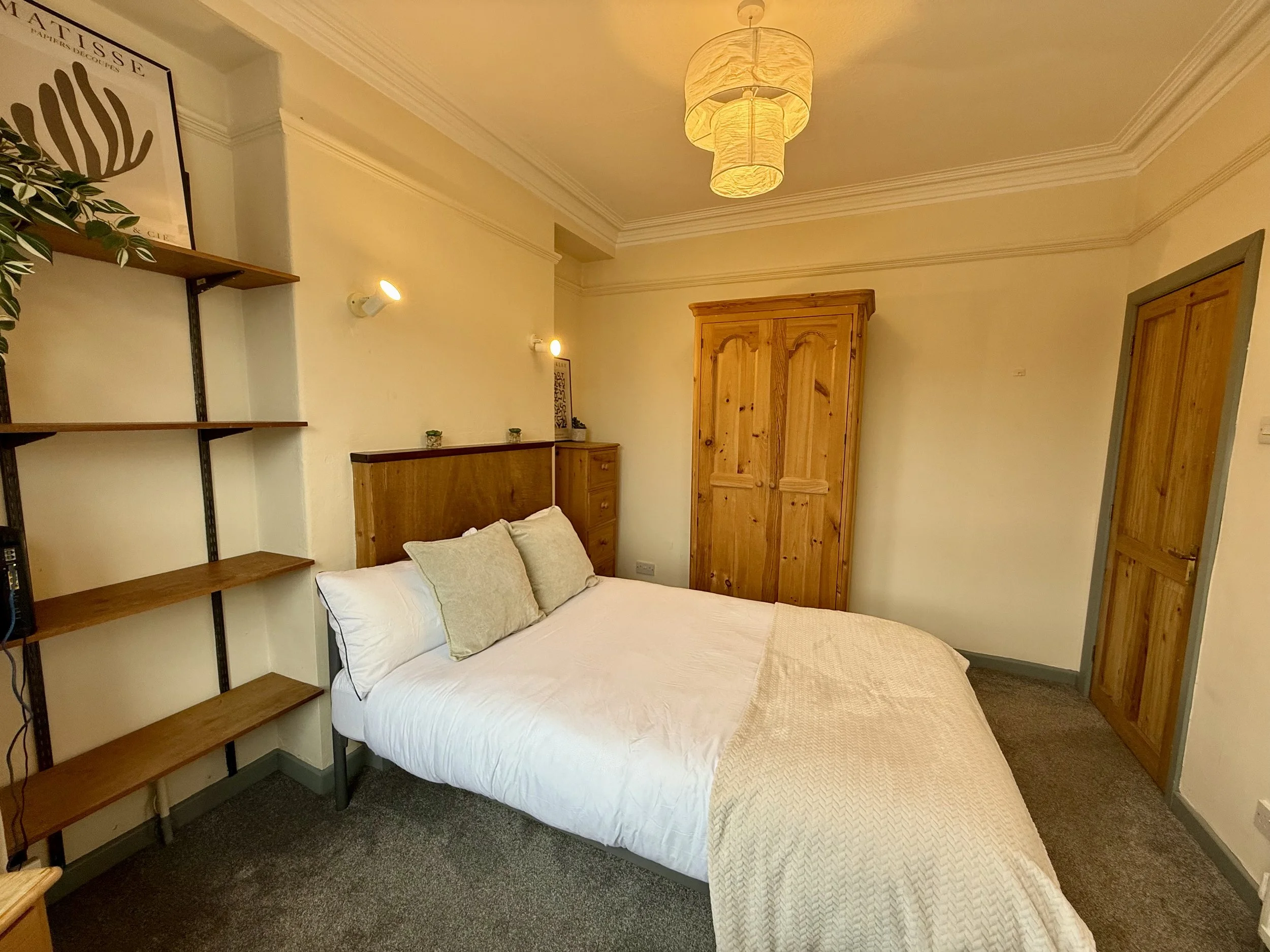 A cozy bedroom with a bed, wooden wardrobe, and a shelf on the wall, illuminated by wall-mounted lights and a ceiling light fixture.