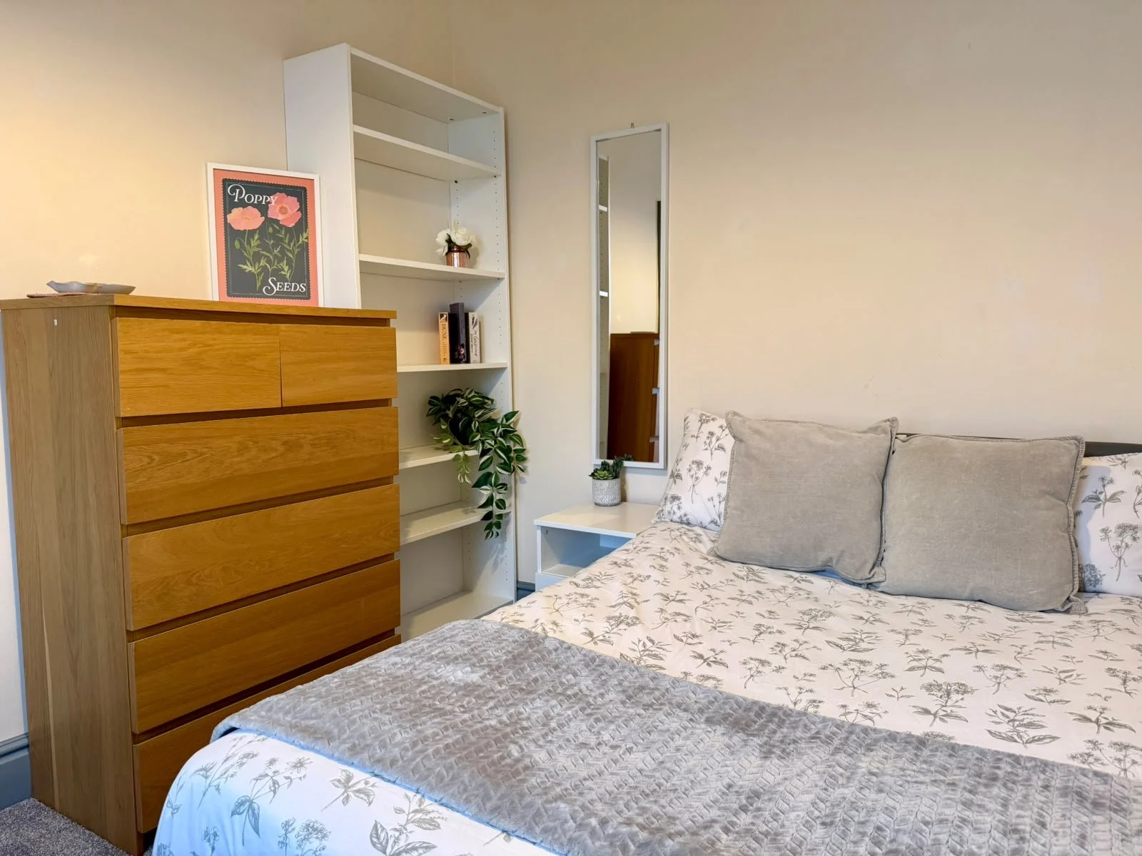 Part of a bedroom with a bed, chest of drawers, and bookshelf, decorated with plants and framed art.