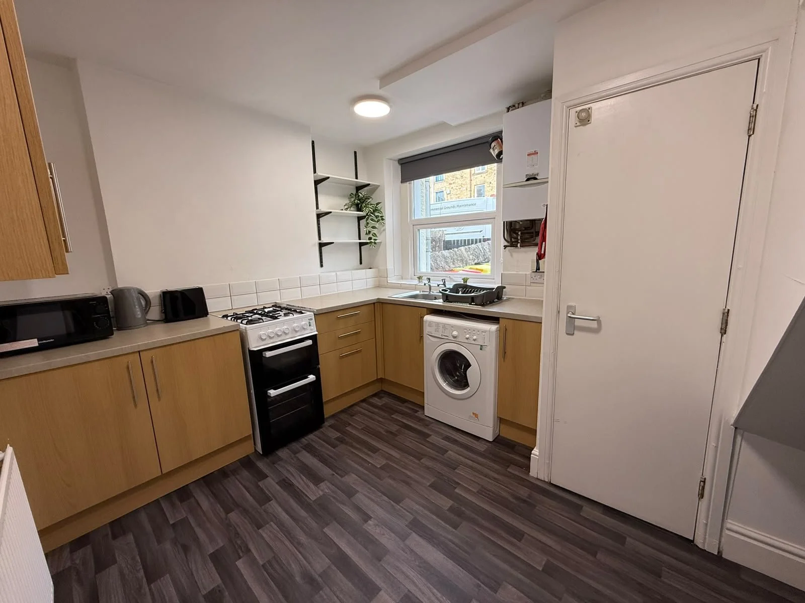 Small kitchen with wooden cabinets, a stove, a washing machine, microwave, kettle, toaster, and a window with a grey roller blind.