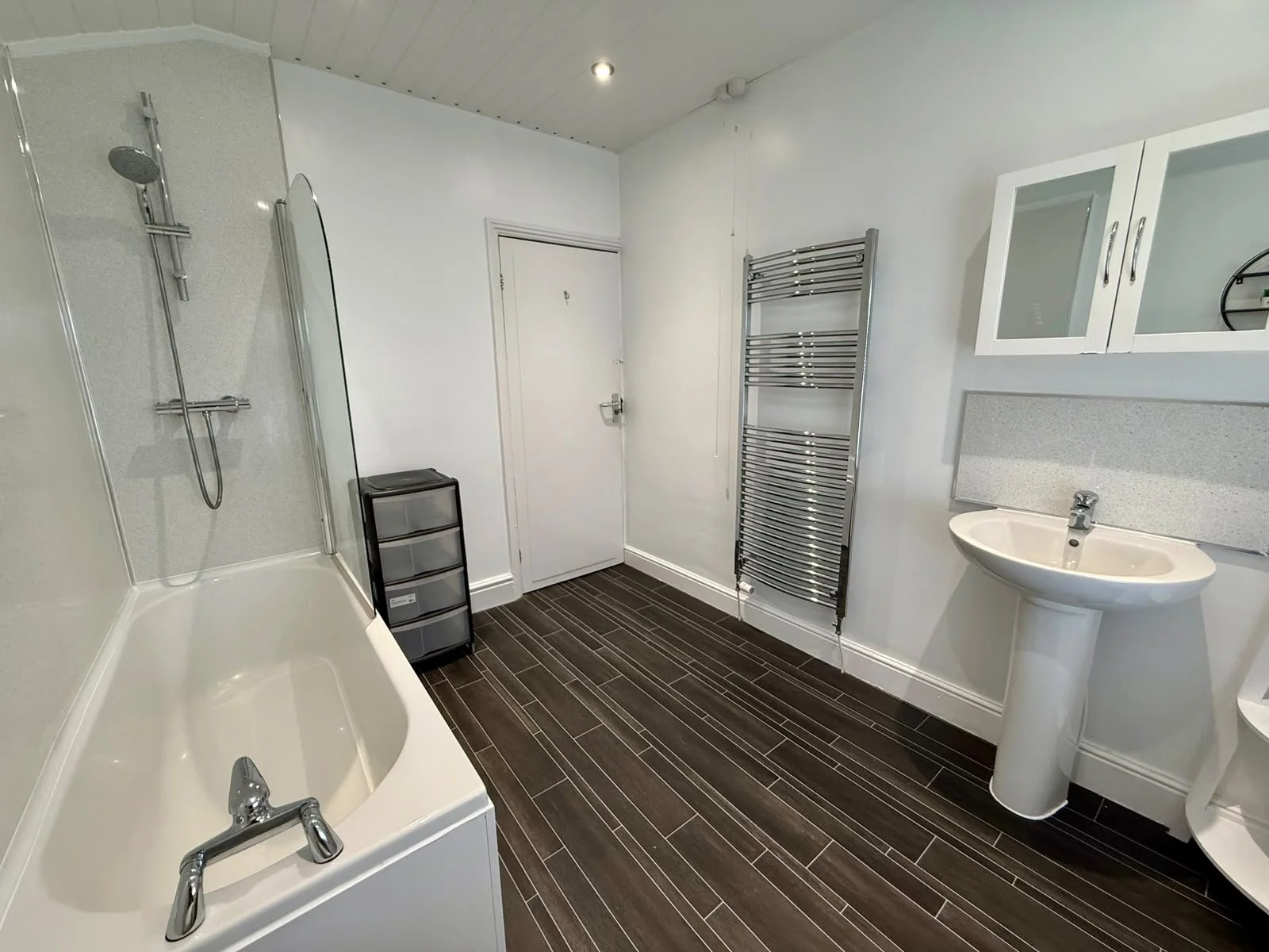A bathroom with a bathtub, a shower area with a glass partition, a small black storage drawer unit, a wall-mounted towel radiator, a white pedestal sink with a mirror cabinet above, and wood-look floor tiles.