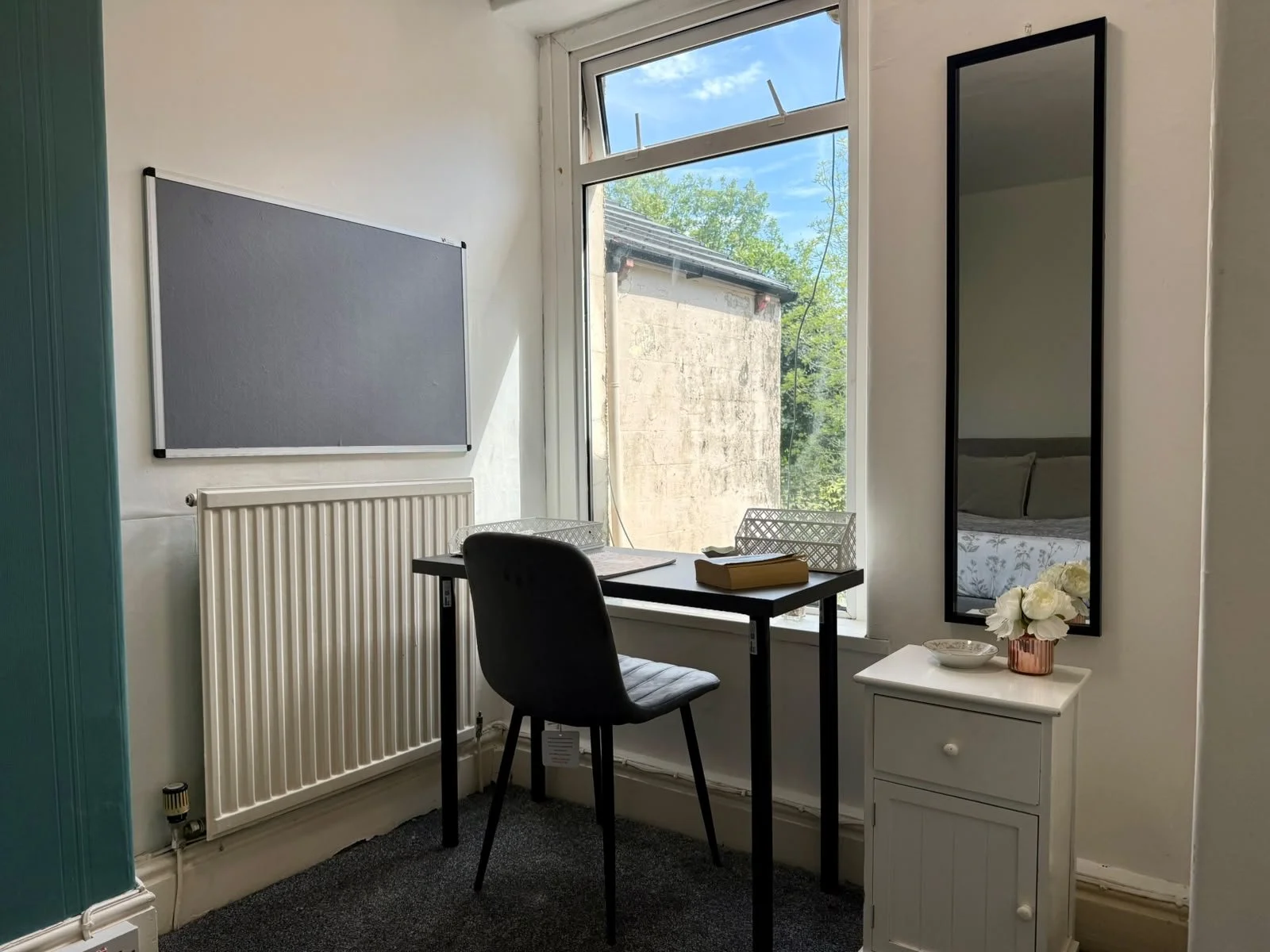 A small home office with a black desk, black chair, and a white side table with a vase of white flowers. There is a large window with a view outside, a black-framed mirror on the wall, and a whiteboard on the wall.