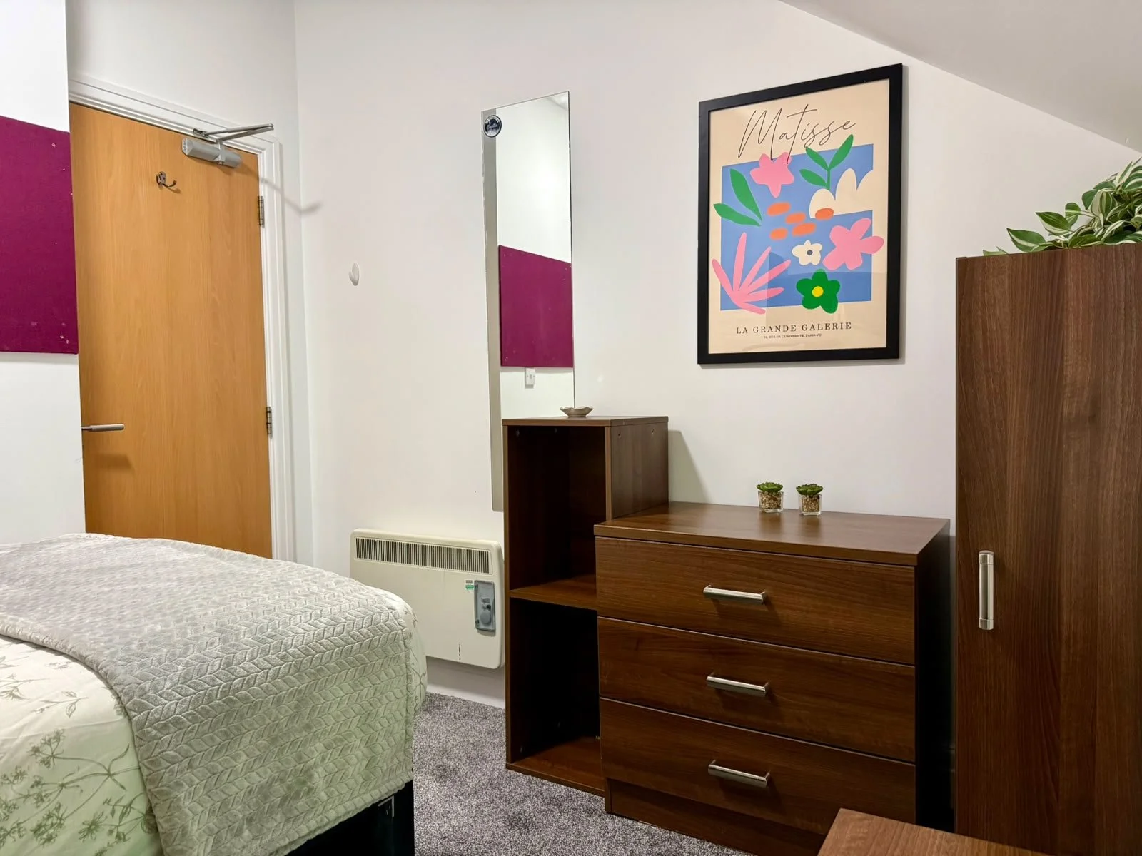 part of a bedroom with a bed, wooden dresser, mirror, wall art, and a door.