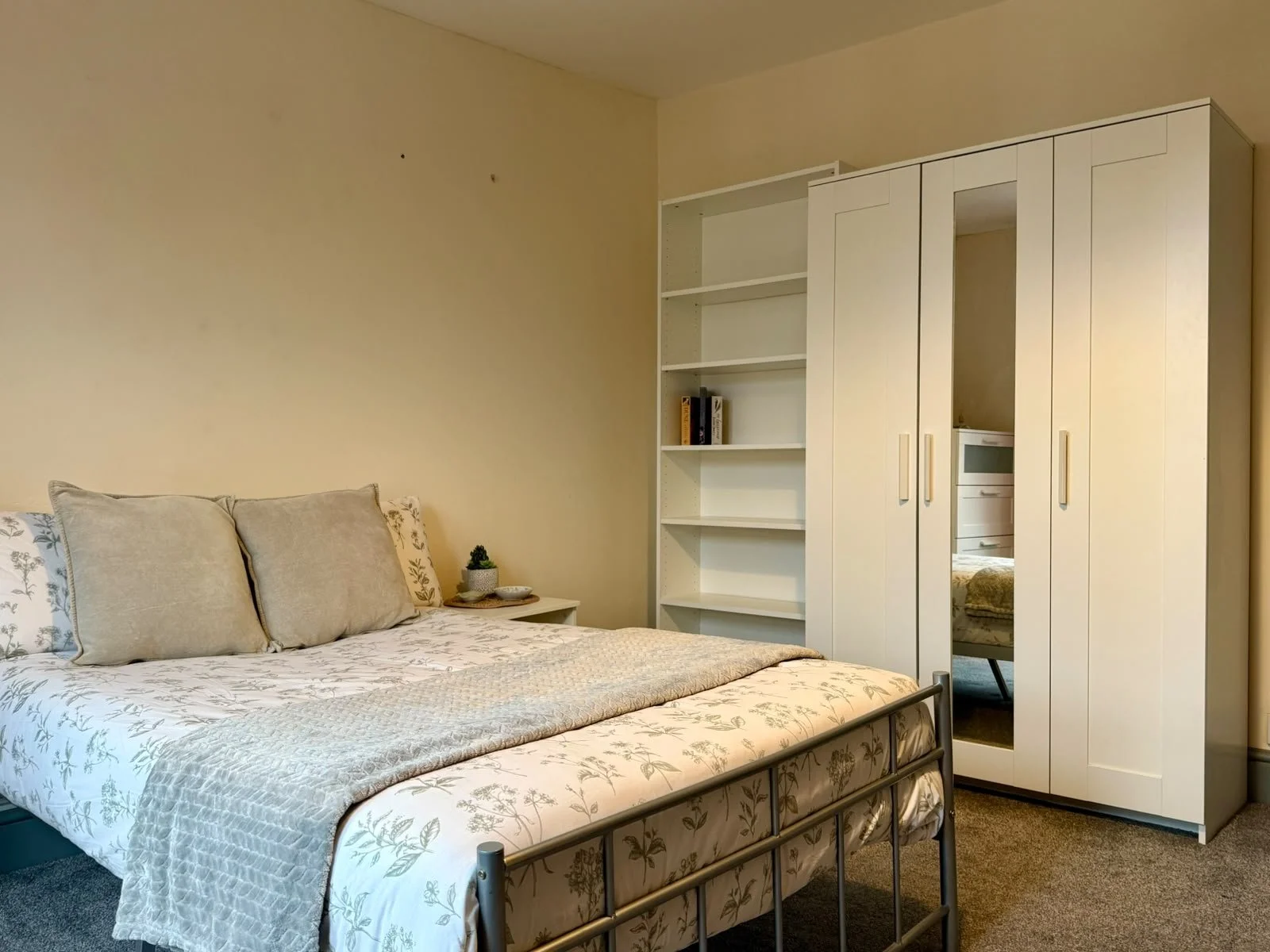 A bedroom with a bed, three pillows, a small nightstand with a potted plant, white open bookshelves, and a white wardrobe with a mirror.