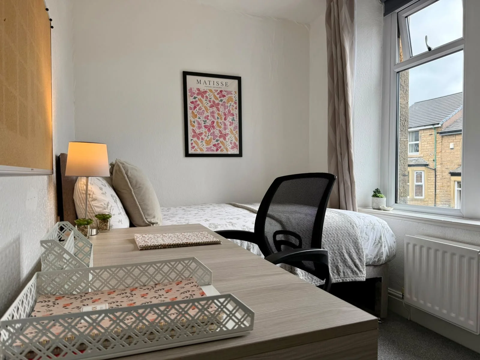 A cozy bedroom with a bed against the wall, decorated with pillows and a bedspread. A bedside table with a lamp and small potted plants. A window with curtains providing natural light. A framed art print titled "Matisse" on the wall. A modern office 