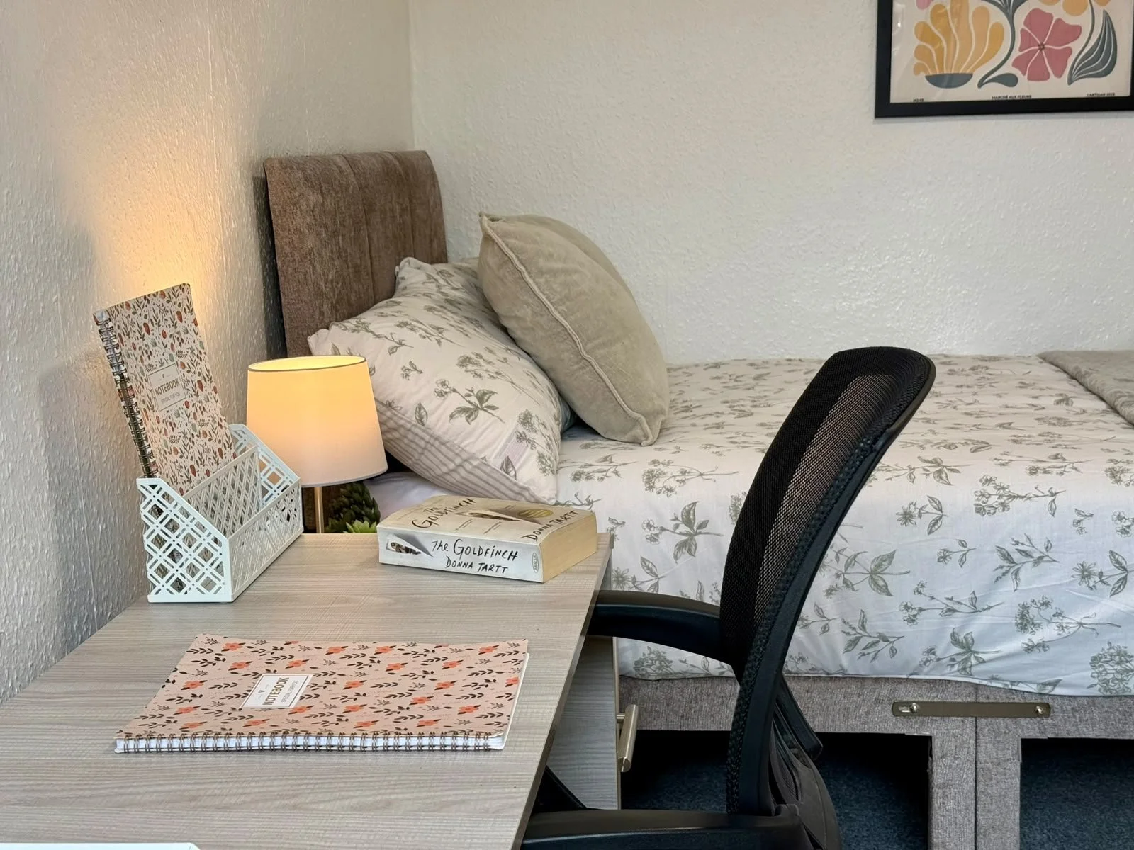 A cozy bedroom with a bed that has floral-patterned bedding and pillows. A wooden nightstand with a table lamp, books, and a notebook. A black ergonomic chair in front of a light wood desk. There is a framed floral artwork on the wall.