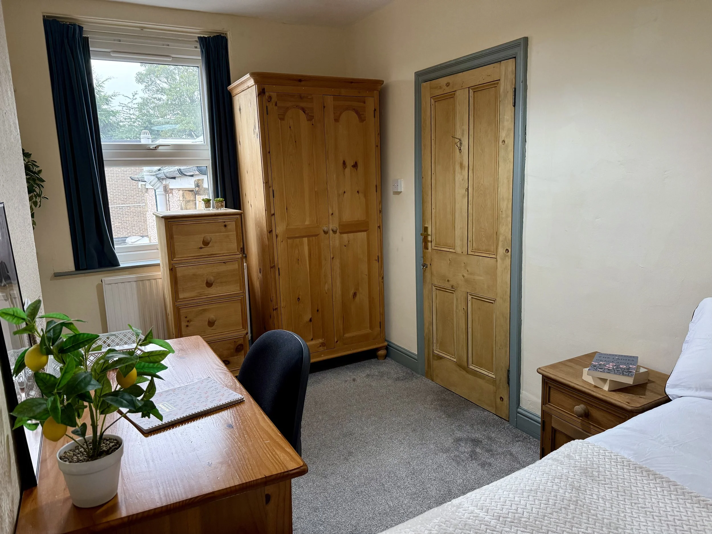 Interior of a bedroom with a wooden table, a potted lemon tree, a bed, a nightstand with a book, a wooden wardrobe, a small chest of drawers, a window with dark curtains, and a wooden door.
