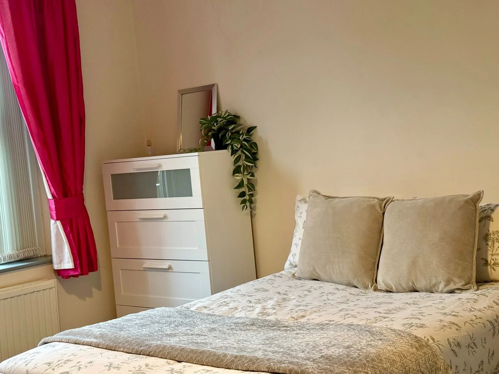 A cozy bedroom with a bed covered in a floral bedspread, beige pillows, a white dresser with glass doors, a mirror, and a green plant next to the dresser. Pink curtains hang over a window with blinds.