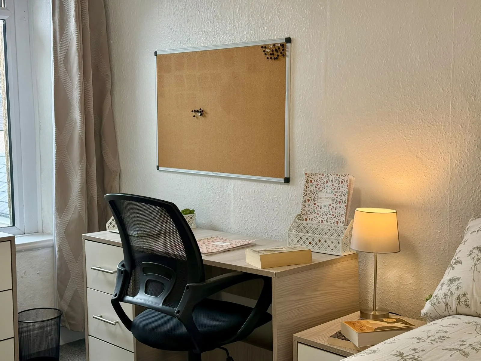 A bedroom corner featuring a desk with a black office chair, a corkboard on the wall, a white desk lamp, a notebook, a small potted plant, and a window with beige curtains.