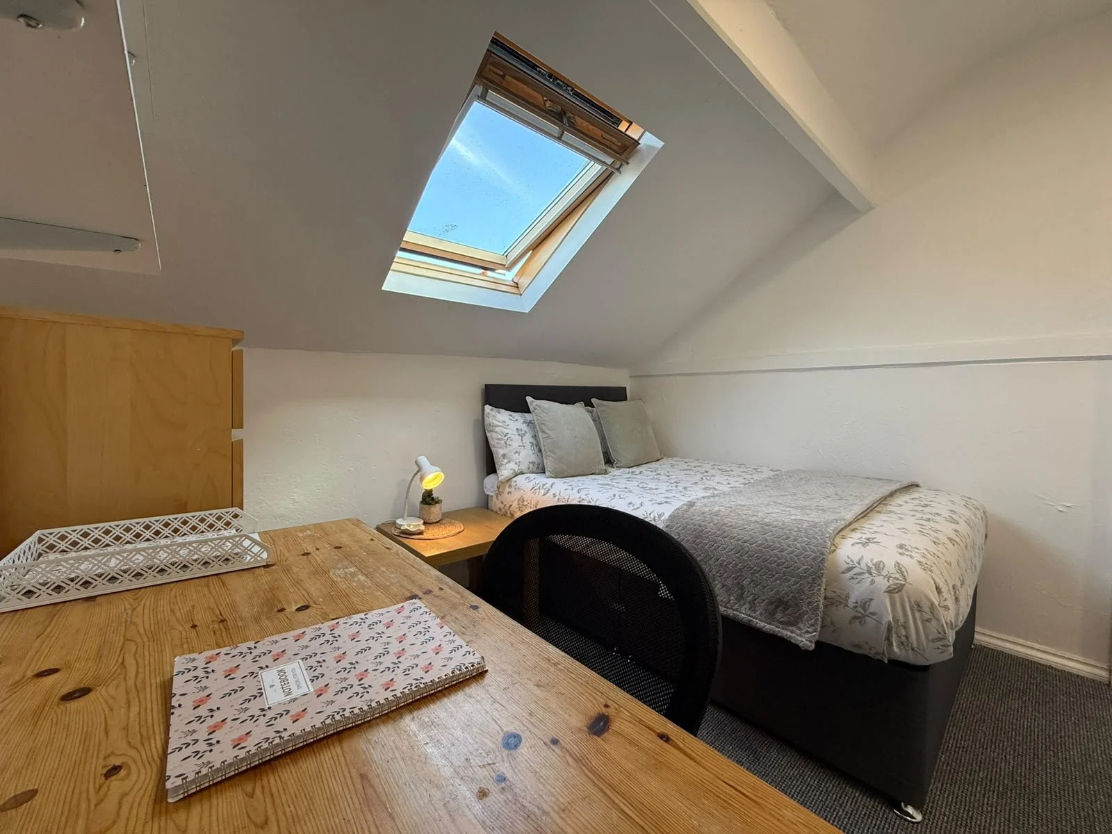A cozy bedroom with a sloped ceiling, featuring a bed with floral and plain pillows, a wooden nightstand with a small lamp and plant, a skylight window showing a blue sky, and a wooden table with a decorative notebook on top.