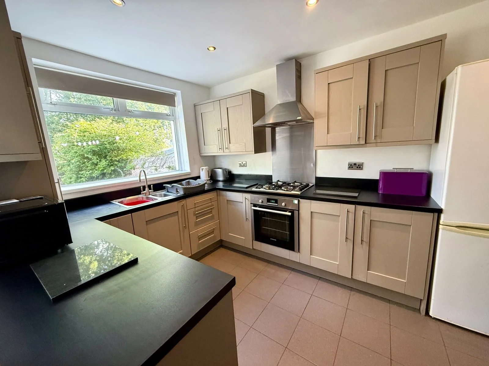 Kitchen with beige cabinets, black countertop, stainless steel oven, gas stove, stainless steel range hood, window with greenery outside, purple toaster, white refrigerator, kettle, toaster, and a black induction cooktop.