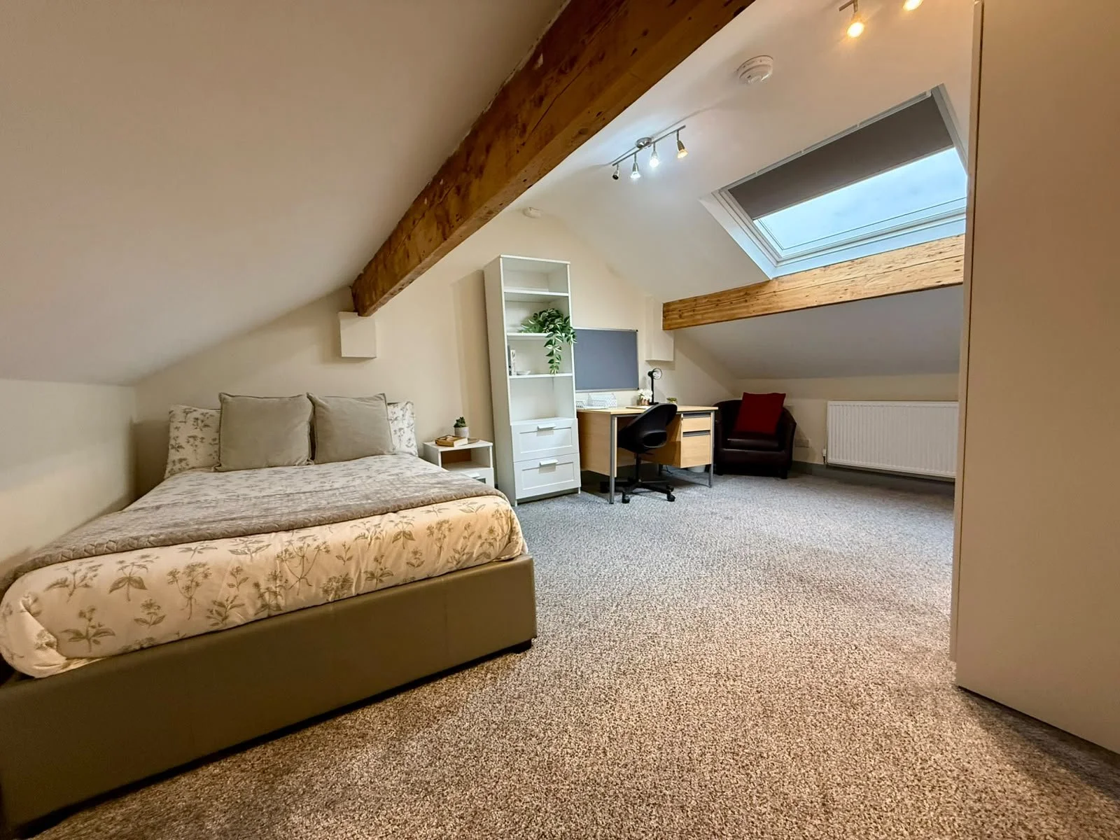 A bedroom with a sloped ceiling, featuring a bed with floral bedding and pillows, a small white nightstand, a white bookshelf, a desk with a computer, a black office chair, a leather armchair with a red pillow, a skylight window, and a ceiling track 