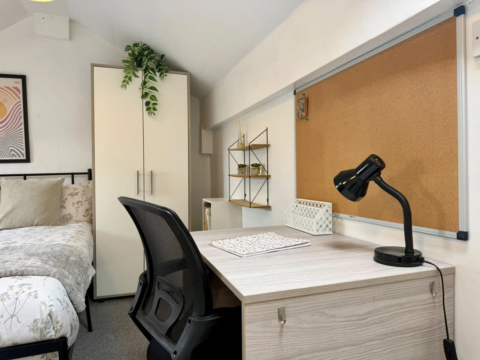 A workspace with a desk, black office chair, desk lamp, corkboard, white storage box, and wall shelves with plants in a bedroom setting.