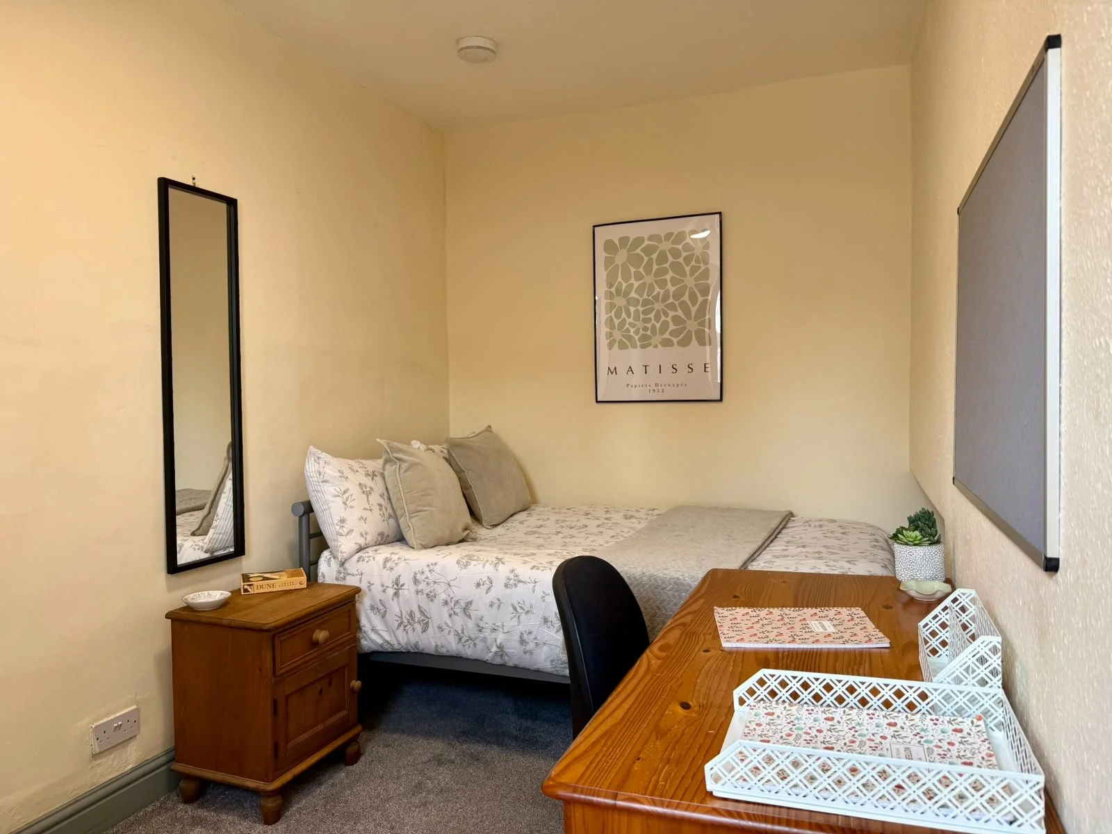 A cozy bedroom with a bed against the wall, beige pillows, and a framed Matisse poster hanging above. There is a wooden nightstand with a small bowl and box of Dune cigarettes. To the right, a wooden table with a white lattice tray and some decorativ